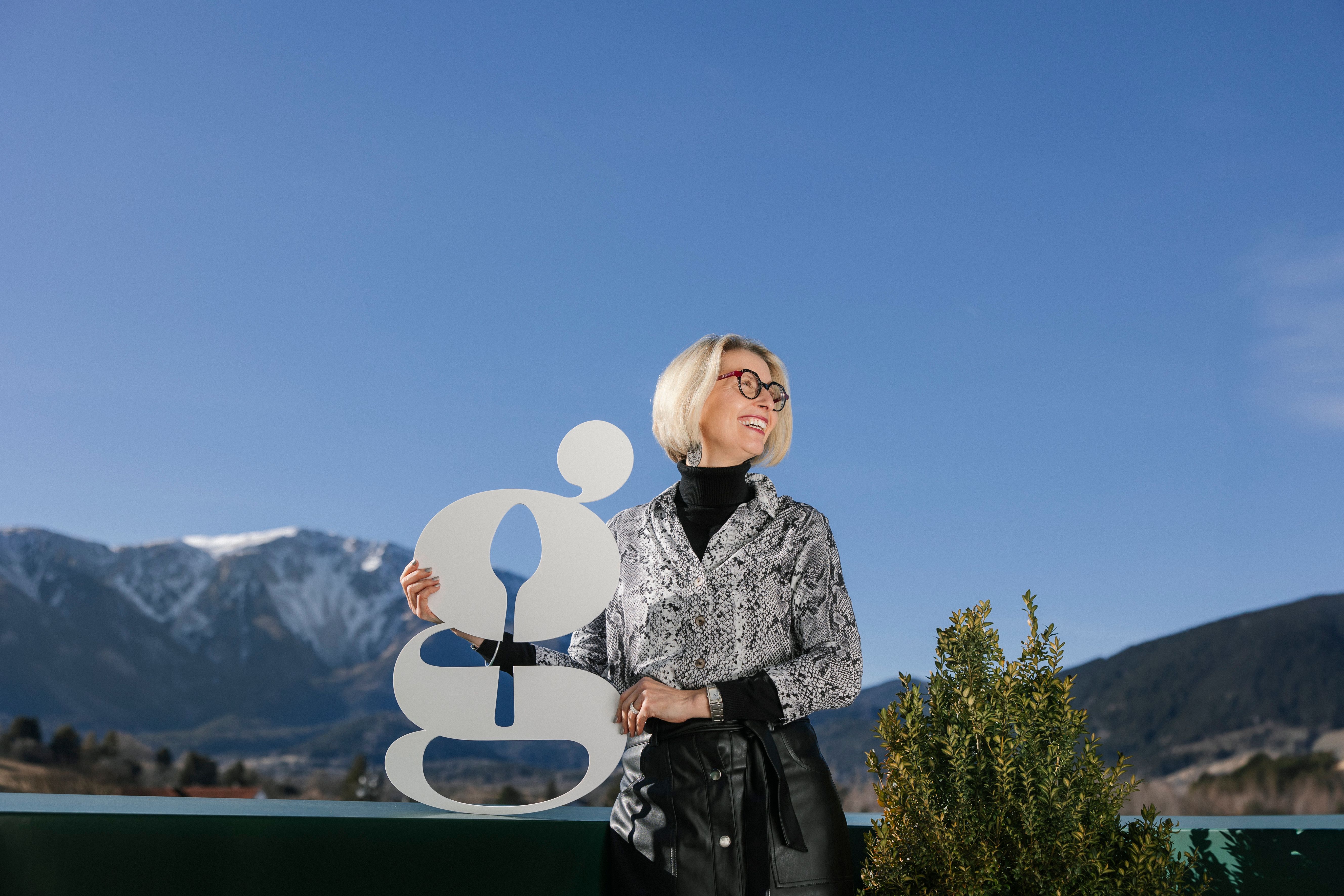 Eine Frau mit blonden Haaren und Brille hält ein großes, stilisiertes 'g' vor einer Berglandschaft.