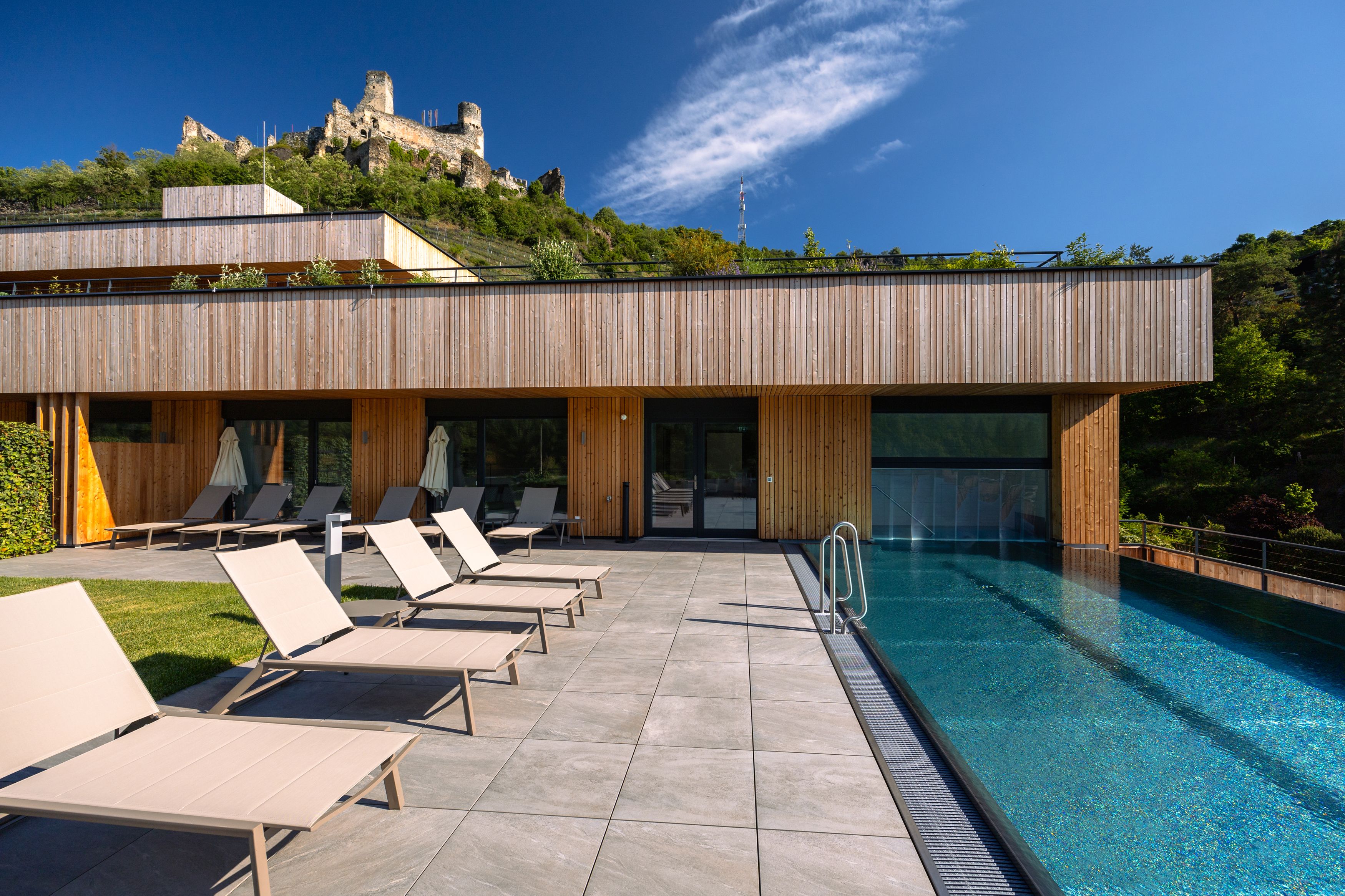 Modernes Hotel mit Pool und Liegestühlen, Burg im Hintergrund.