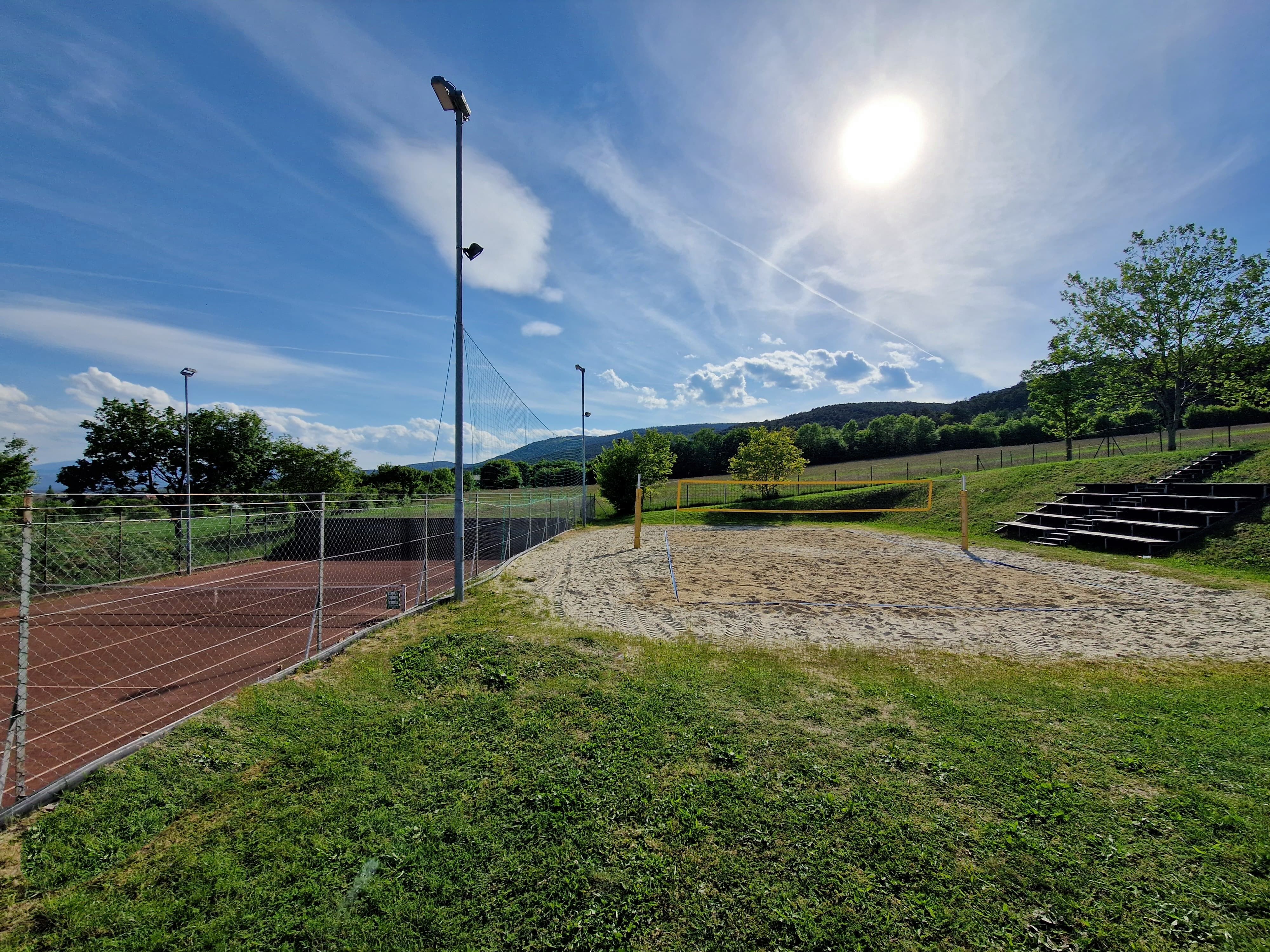 Ein sonniger Volleyballplatz mit Sand und einem Netz, umgeben von grüner Landschaft und Bäumen.