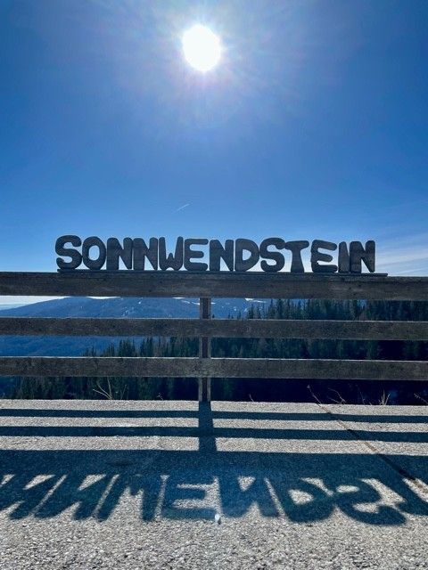 Großbuchstaben mit 'Sonnwendstein' vor sonnigem Himmel und Berglandschaft.