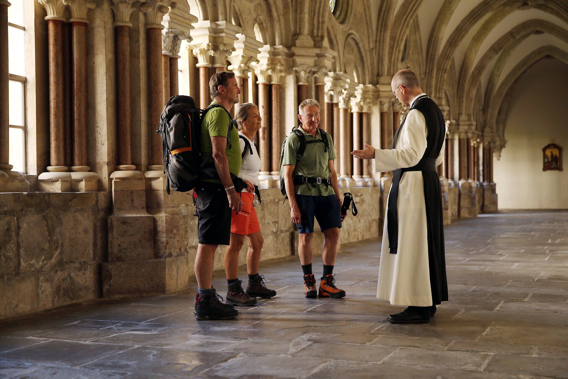 In der beeindruckenden Klosterarchitektur des Stifts Lilienfeld treffen sich Pilger, um ihre Reise zu teilen und neue Kraft zu schöpfen. Die Atmosphäre ist erfüllt von der Stille des Ortes, während die sanften Lichtstrahlen durch die hohen Fenster fallen und die historischen Mauern zum Leben erwecken.