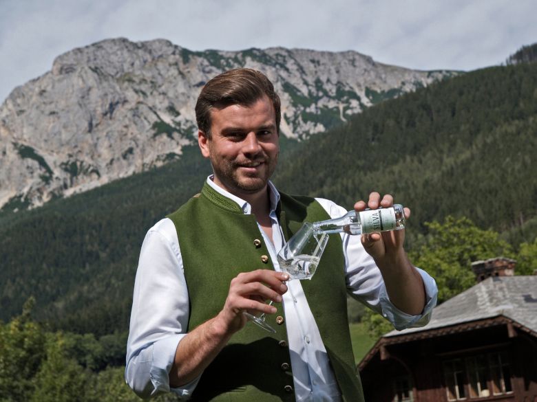 Mann in traditioneller Kleidung schenkt ein Getränk in ein Glas ein, mit Berglandschaft im Hintergrund.