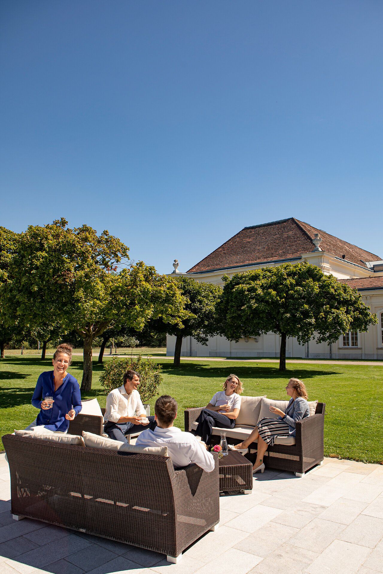 Inmitten einer malerischen Gartenlandschaft genießen die Gäste entspannte Gespräche und erfrischende Getränke. Die strahlende Sonne und der klare Himmel schaffen eine einladende Atmosphäre, die zum Verweilen einlädt. Umgeben von üppigem Grün und historischen Gebäuden wird der Sommer hier zu einem unvergesslichen Erlebnis.