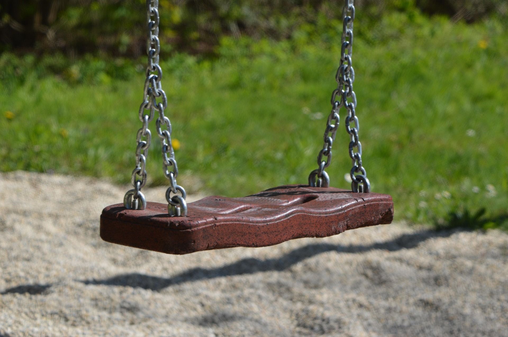 Eine leere Schaukel aus Holz und Metallketten hängt über einem Kiesboden, umgeben von grünem Gras.