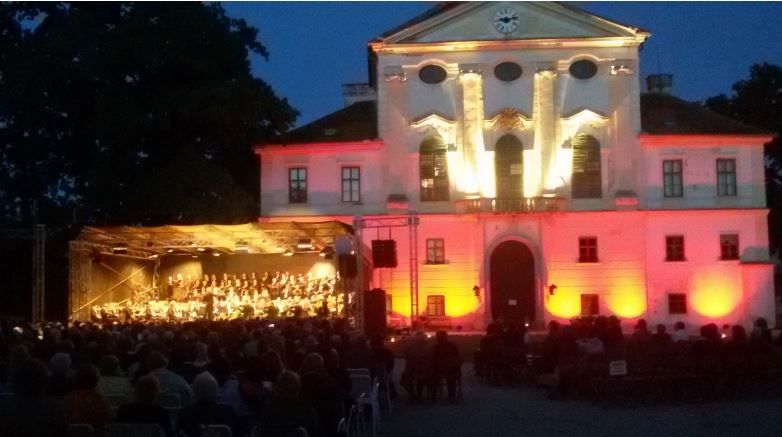 Ein beleuchtetes Gebäude mit einem Orchester, das auf einer Bühne im Freien spielt, umgeben von einem sitzenden Publikum bei Nacht.