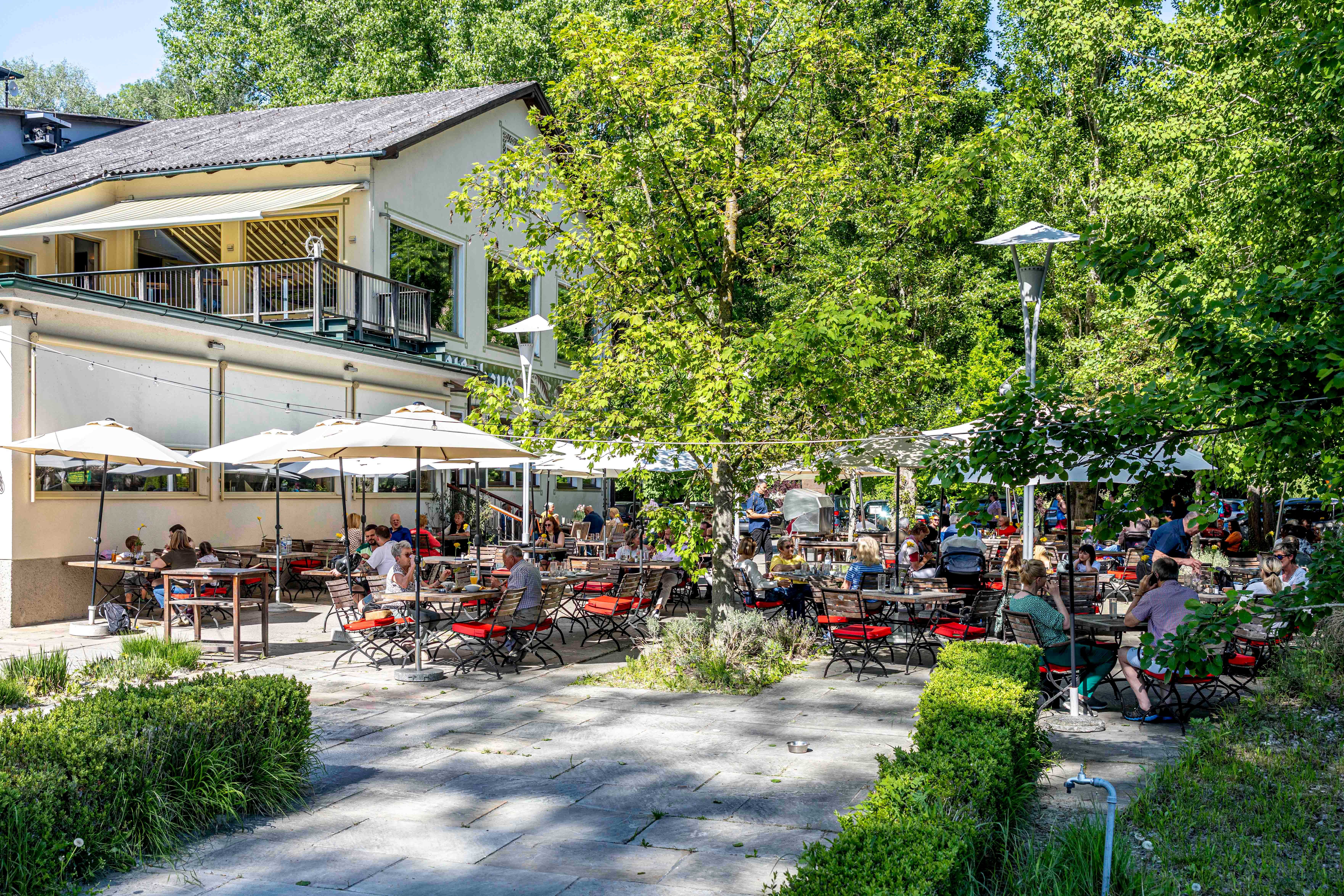 Ein Gastgarten mit vielen Menschen, die an Tischen unter Sonnenschirmen sitzen, umgeben von Bäumen und einem Gebäude im Hintergrund.