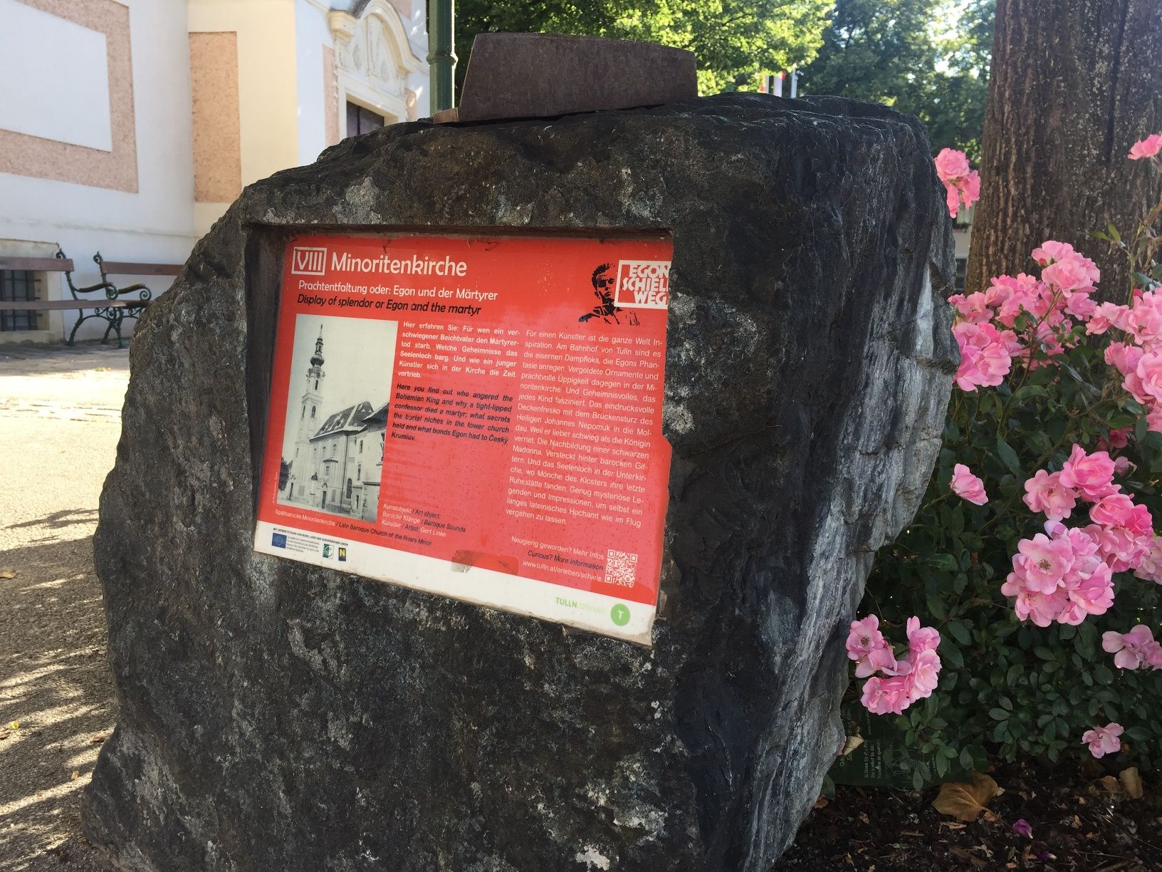 Steintafel mit Informationen zur Minoritenkirche, umgeben von Blumen.