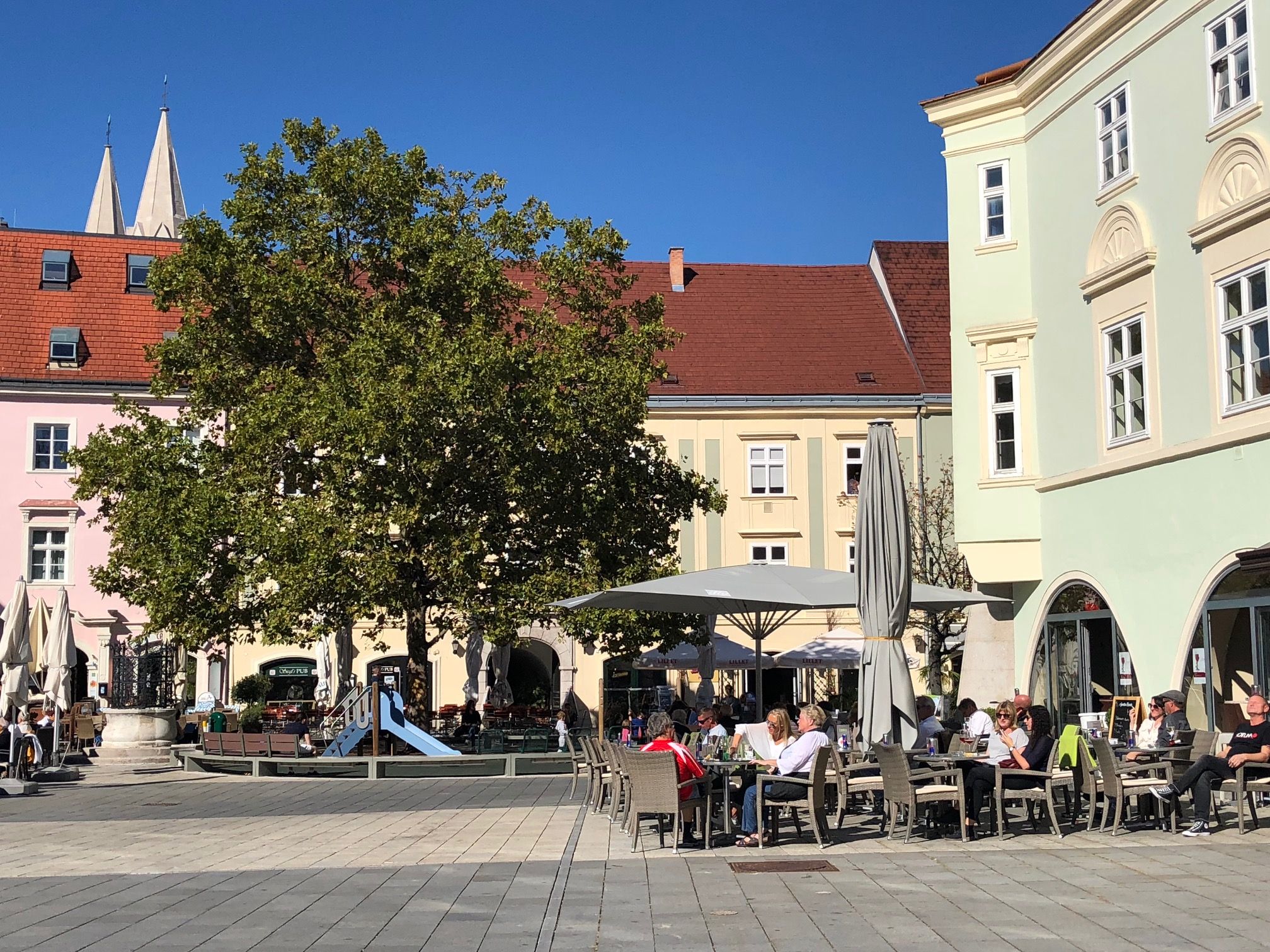 Sonniger Hauptplatz mit einem Baum und Spielplatz beim Eterno. Menschen sitzen draußen und genießen das Wetter.