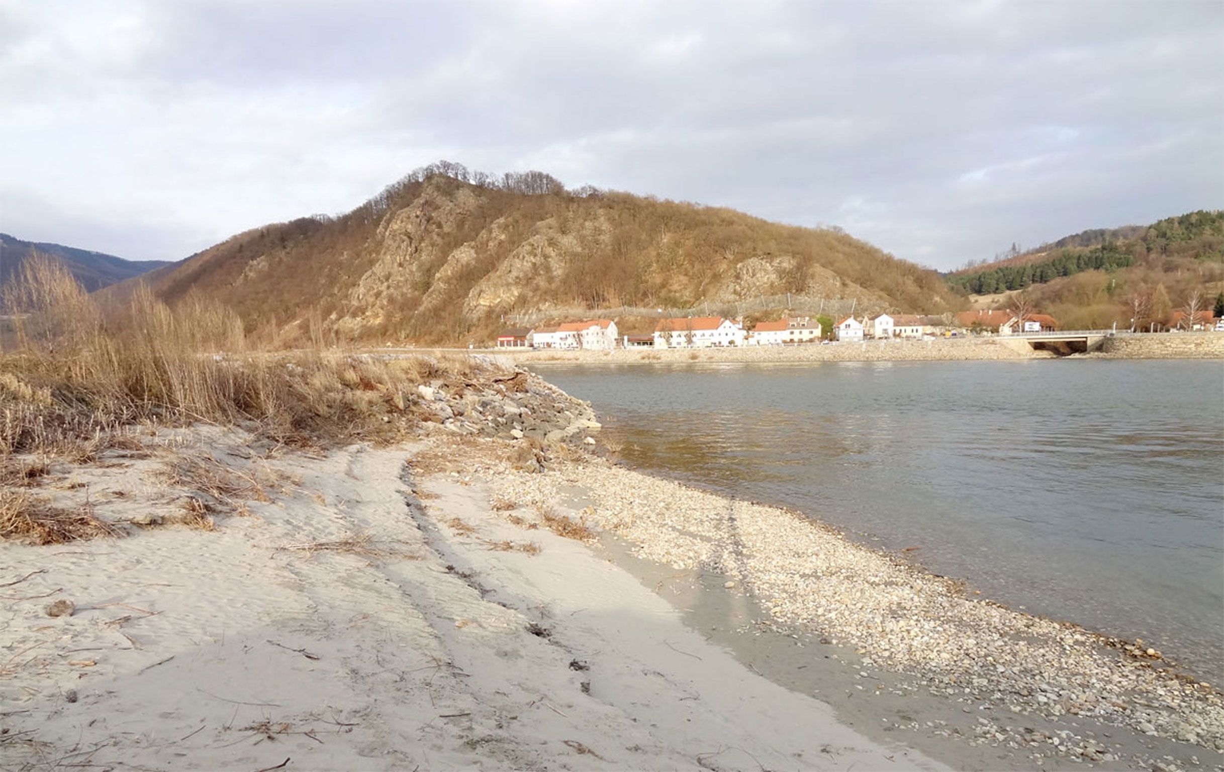 Ein ruhiger Strand mit Kieselsteinen, im Hintergrund ein Dorf und bewaldete Hügel.