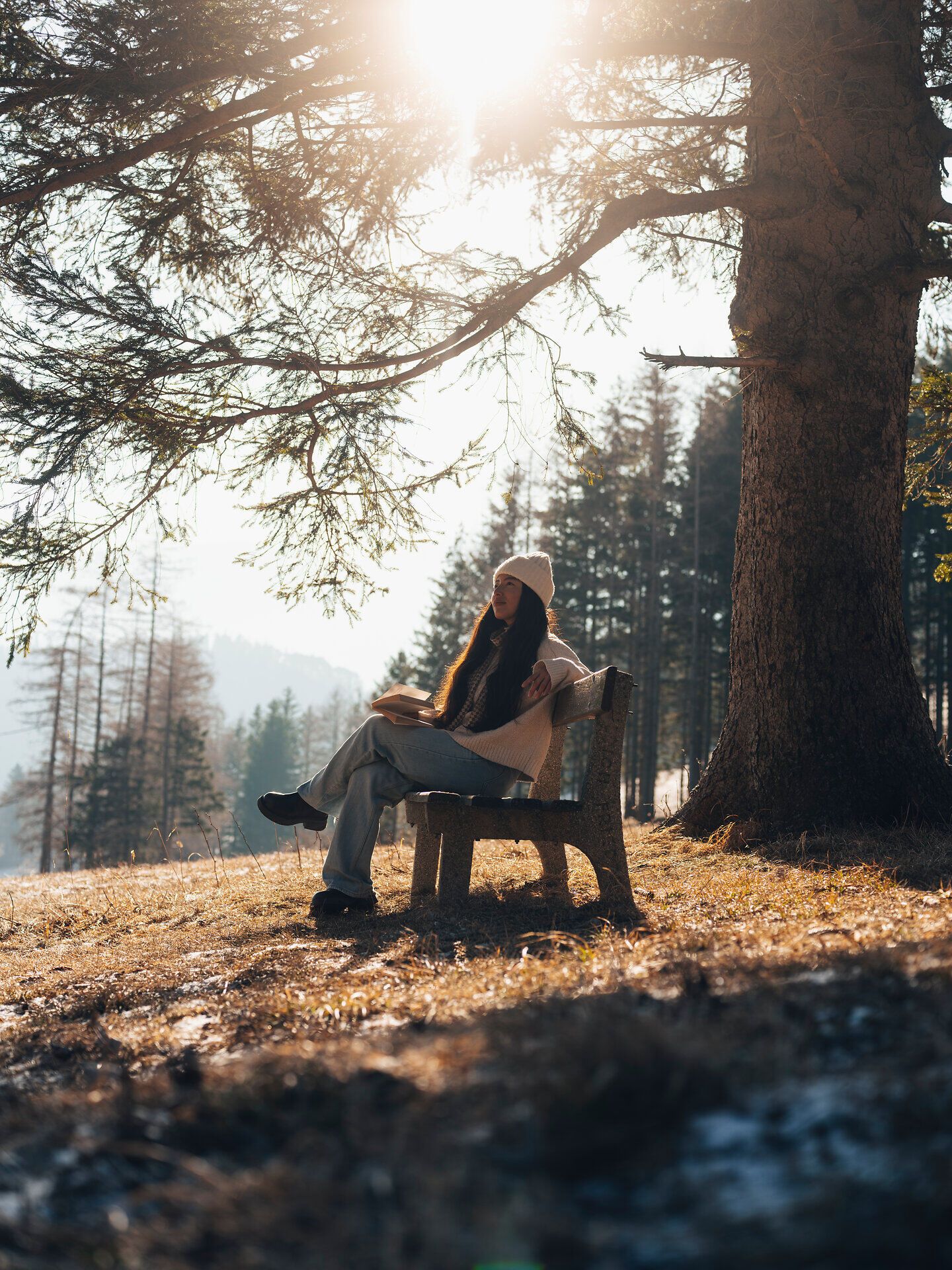 In der winterlichen Landschaft der Wiener Alpen sitzt eine junge Frau auf einer rustikalen Bank, umgeben von schneebedeckten Bäumen und sanften Hügeln. Die warmen Sonnenstrahlen brechen durch die Äste und schaffen eine friedliche Atmosphäre, die zur Achtsamkeit einlädt. Hier, in der Stille der Natur, wird der Winter zu einem Ort der Besinnung und Erholung.