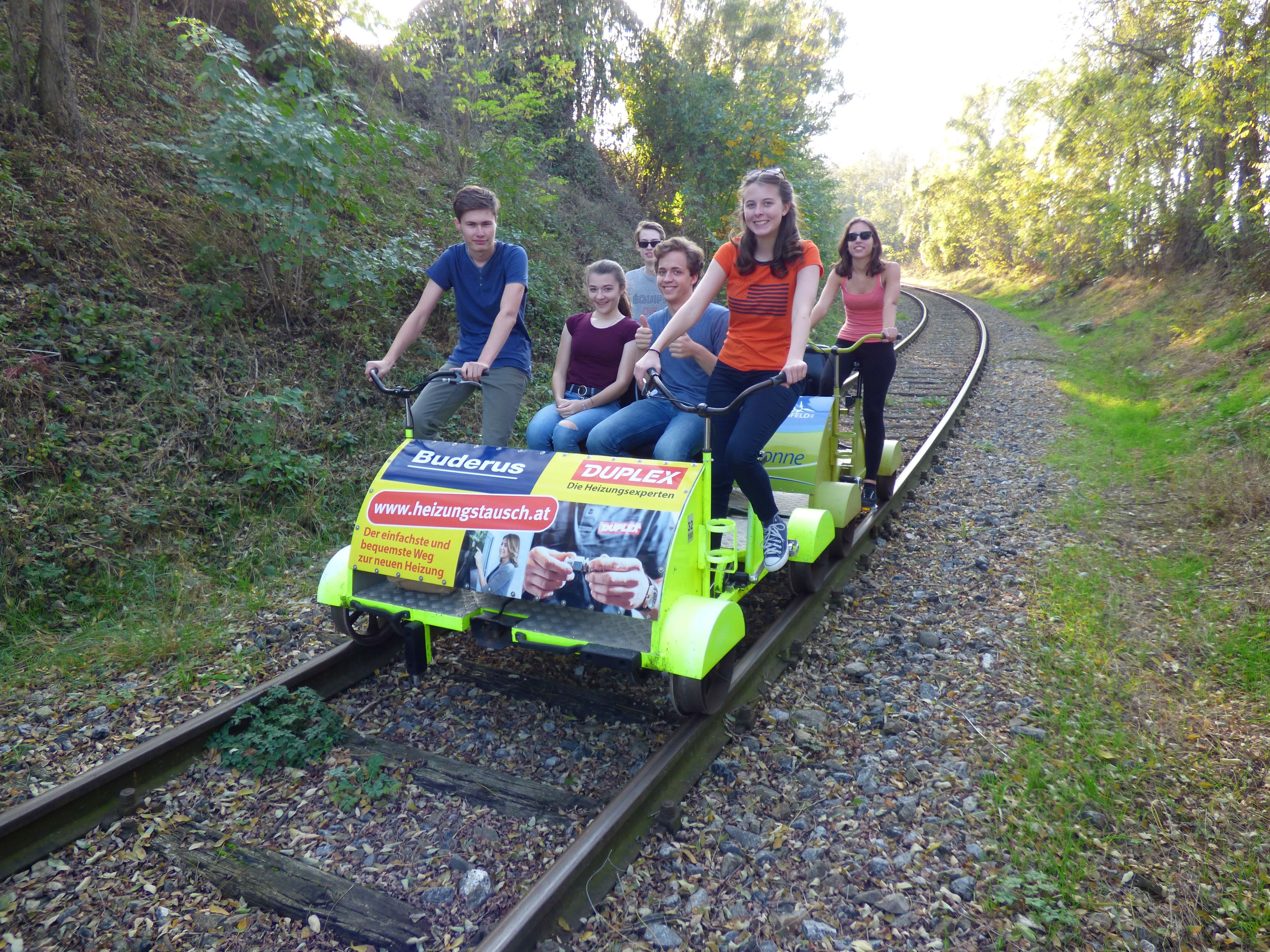 Gruppe von Menschen fährt auf einer Draisine auf Bahngleisen durch eine bewaldete Landschaft.