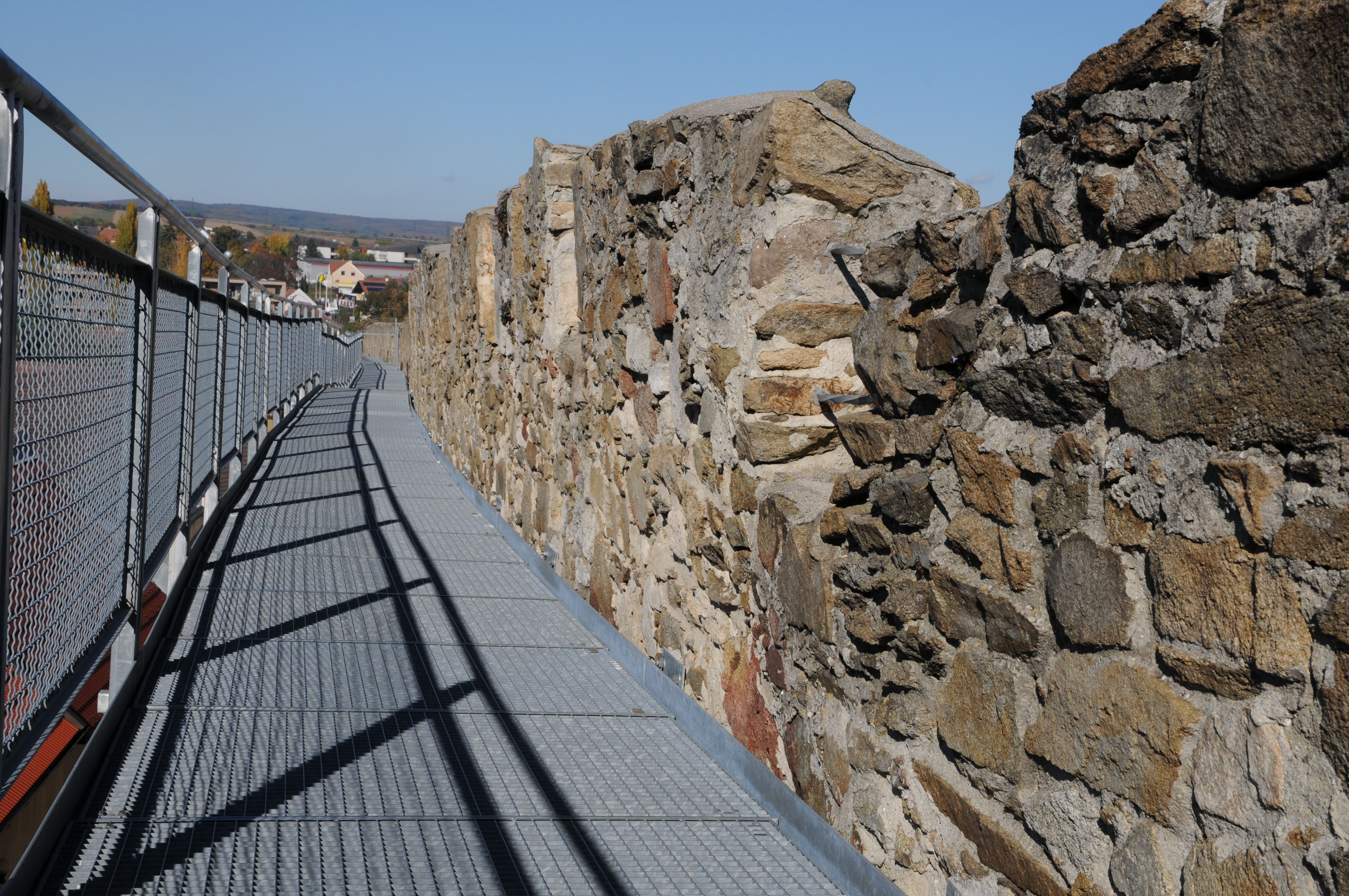 Ein Metallsteg entlang einer alten Steinmauer mit Blick auf eine Stadt im Hintergrund.