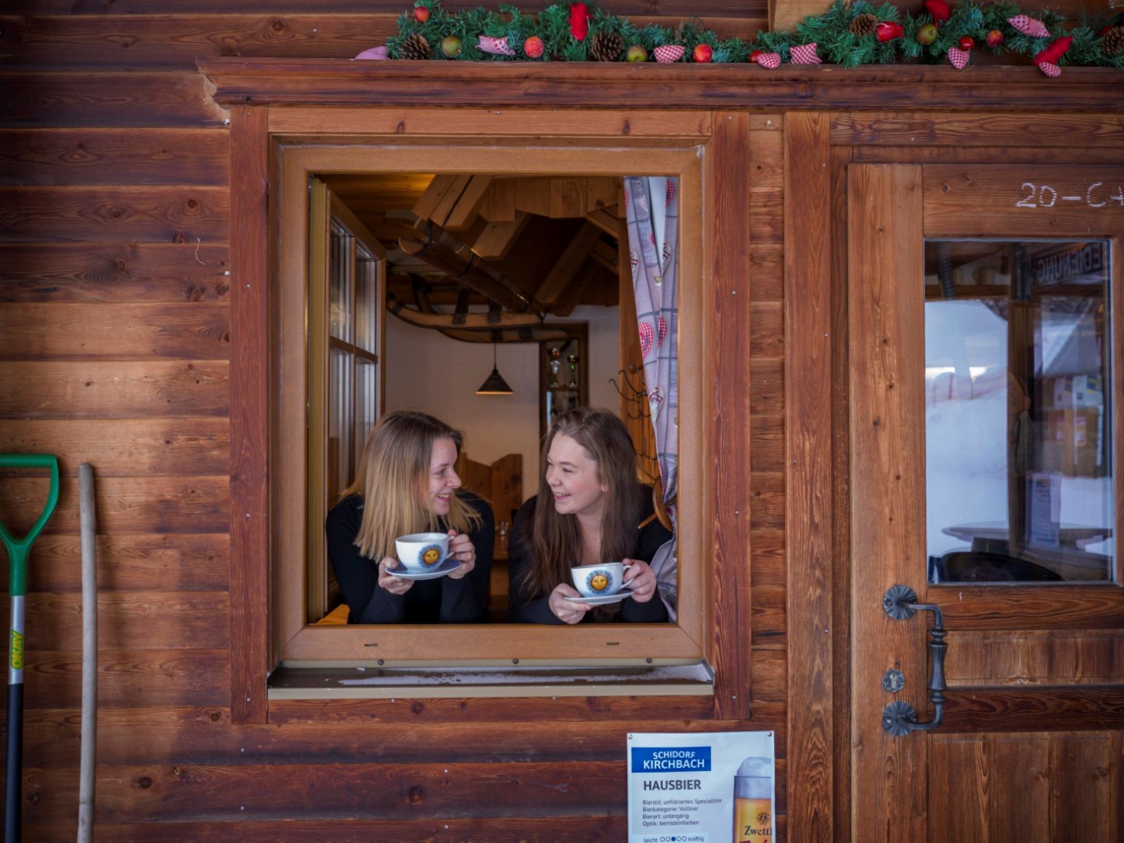 Zwei Frauen schauen aus einem Fenster einer Holzhütte und halten Tassen in den Händen.