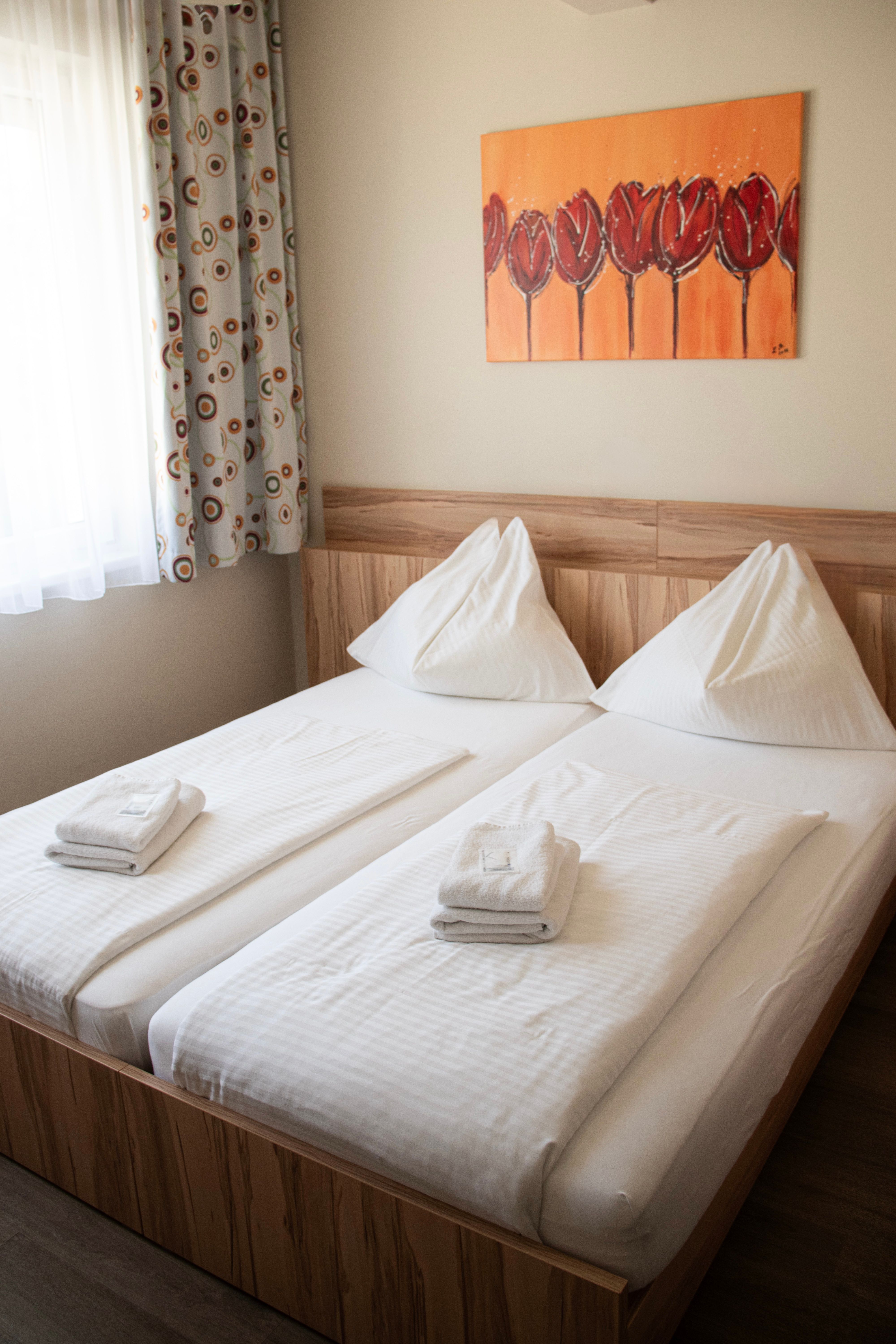 Doppelbett im Kolpinghaus mit weißer Bettwäsche, Handtüchern, Fenster mit gemusterten Vorhängen und Wandbild mit roten Tulpen.