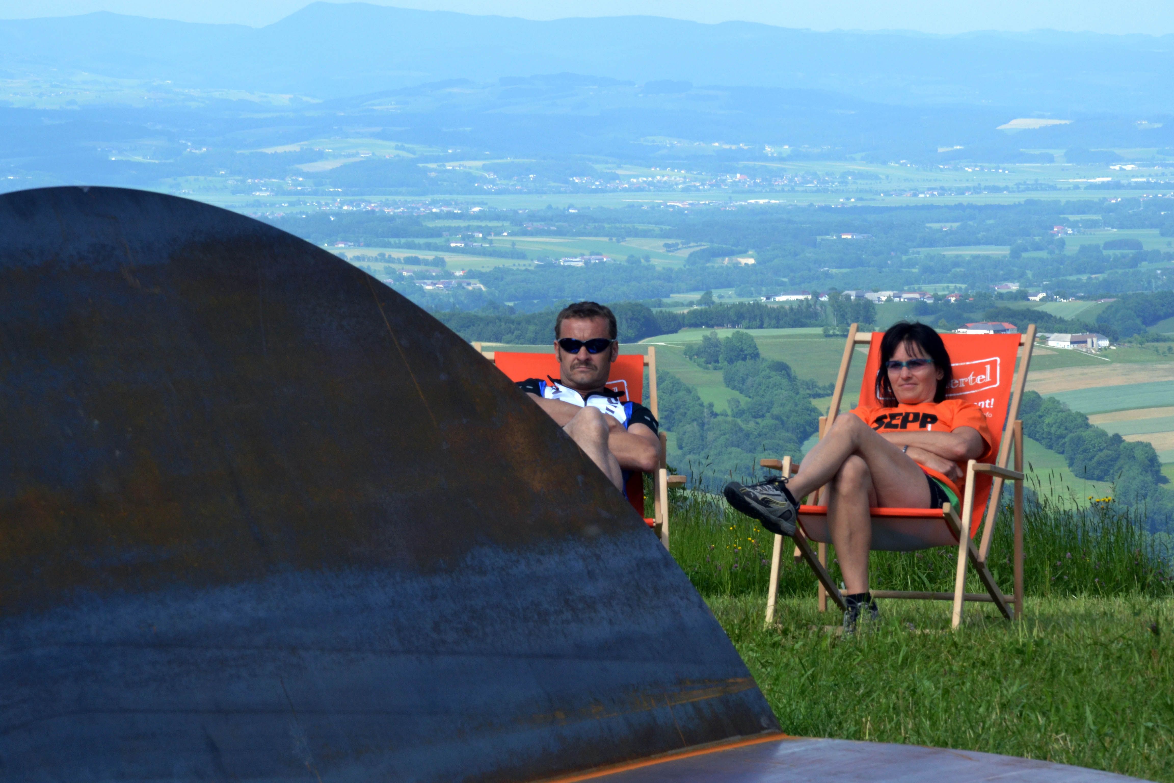 Zwei Personen entspannen in Liegestühlen auf einer Wiese mit Blick auf eine weite Landschaft.