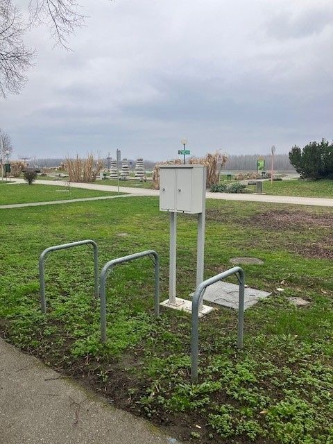 E-Bike Ladestation auf einer Wiese mit Fahrradständern und einem Weg im Hintergrund.