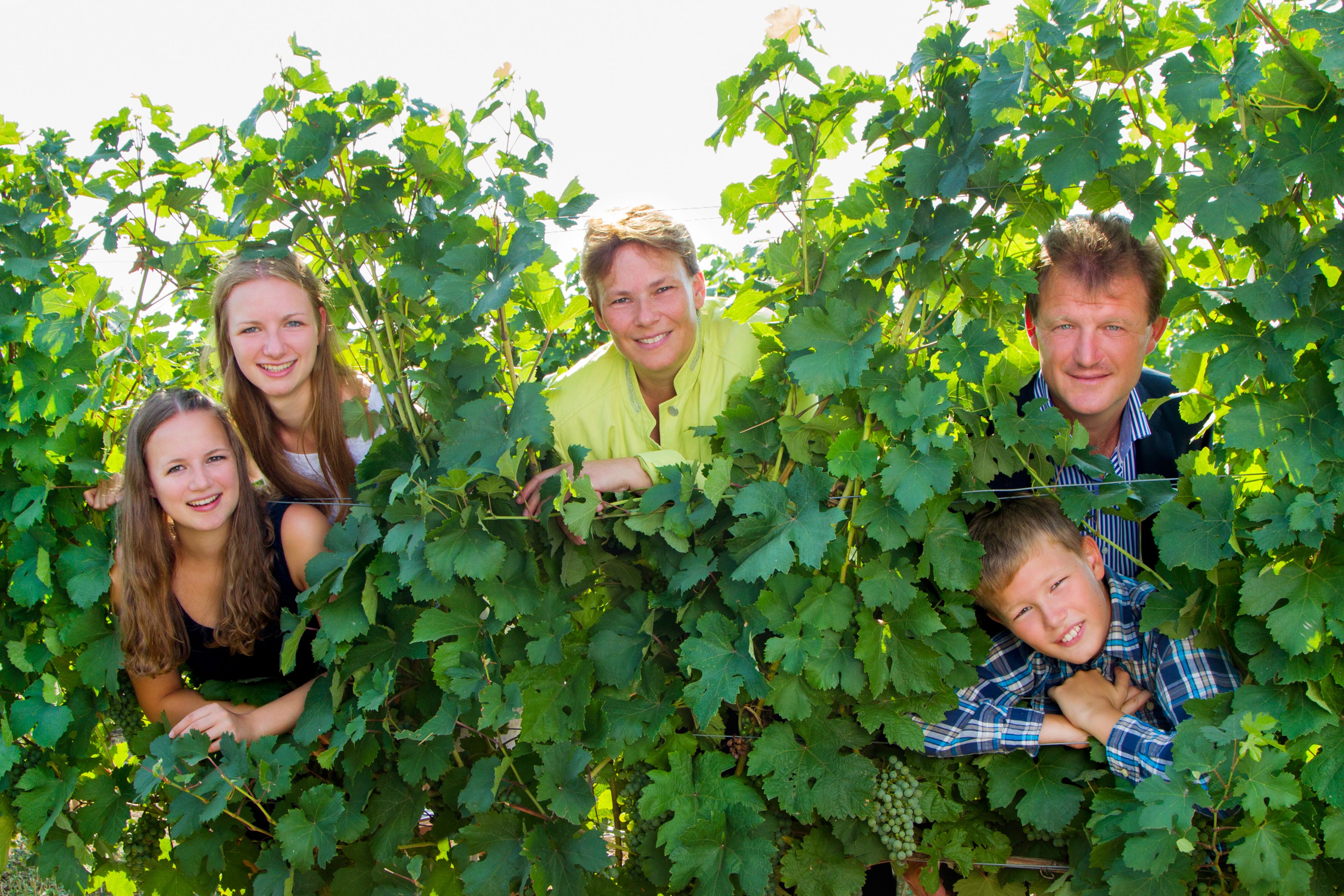 Eine Familie schaut lächelnd durch Weinreben hindurch.