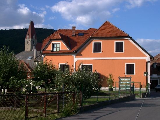 Ein orangefarbenes Haus mit roten Dachziegeln und einem Kirchturm im Hintergrund.
