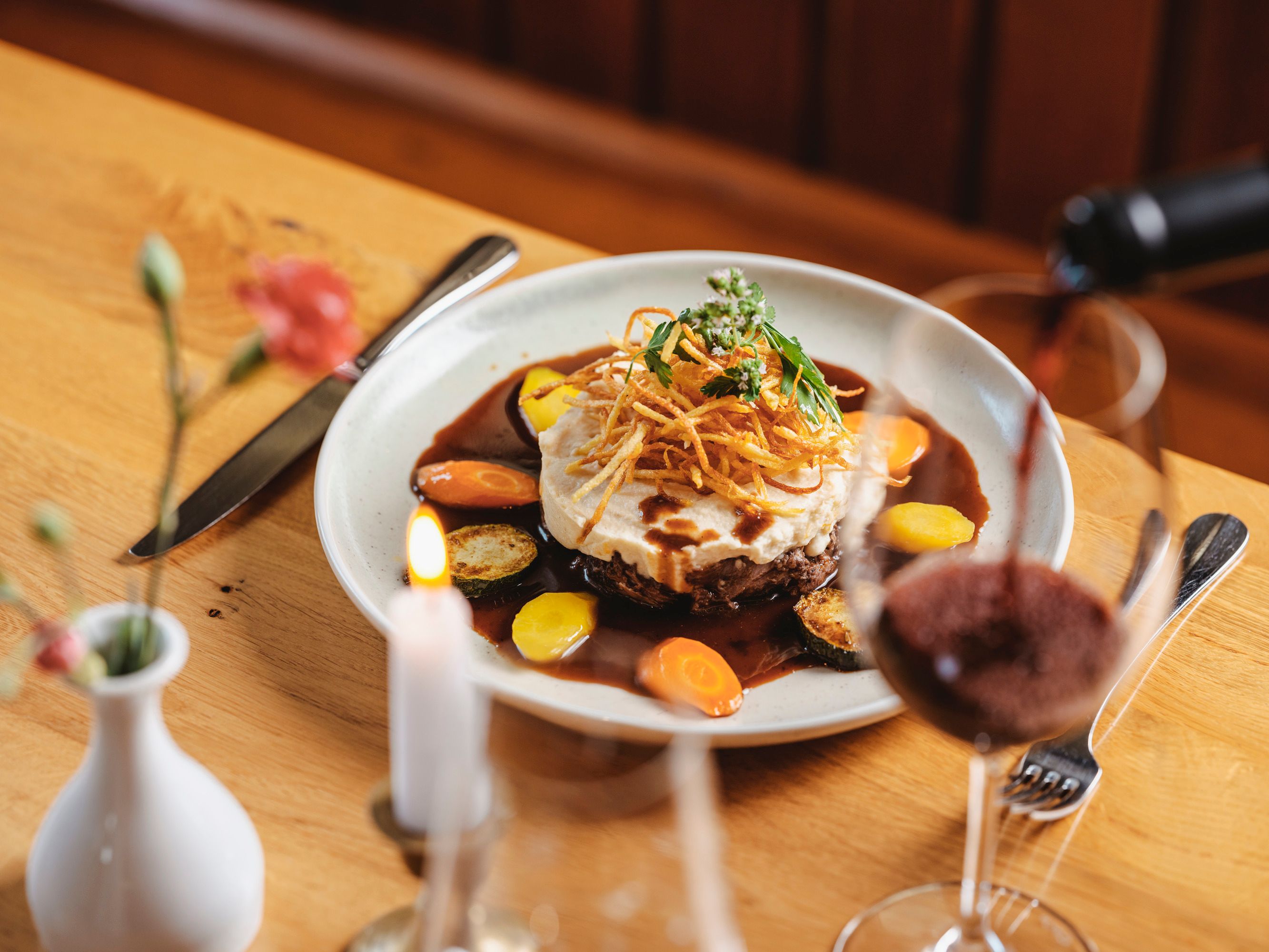 Ein Teller mit Ochsenschlepp in Zweigeltsauce, garniert mit Erdäpfelpüree und Gemüse, auf einem Holztisch. Ein Glas Rotwein wird eingeschenkt.