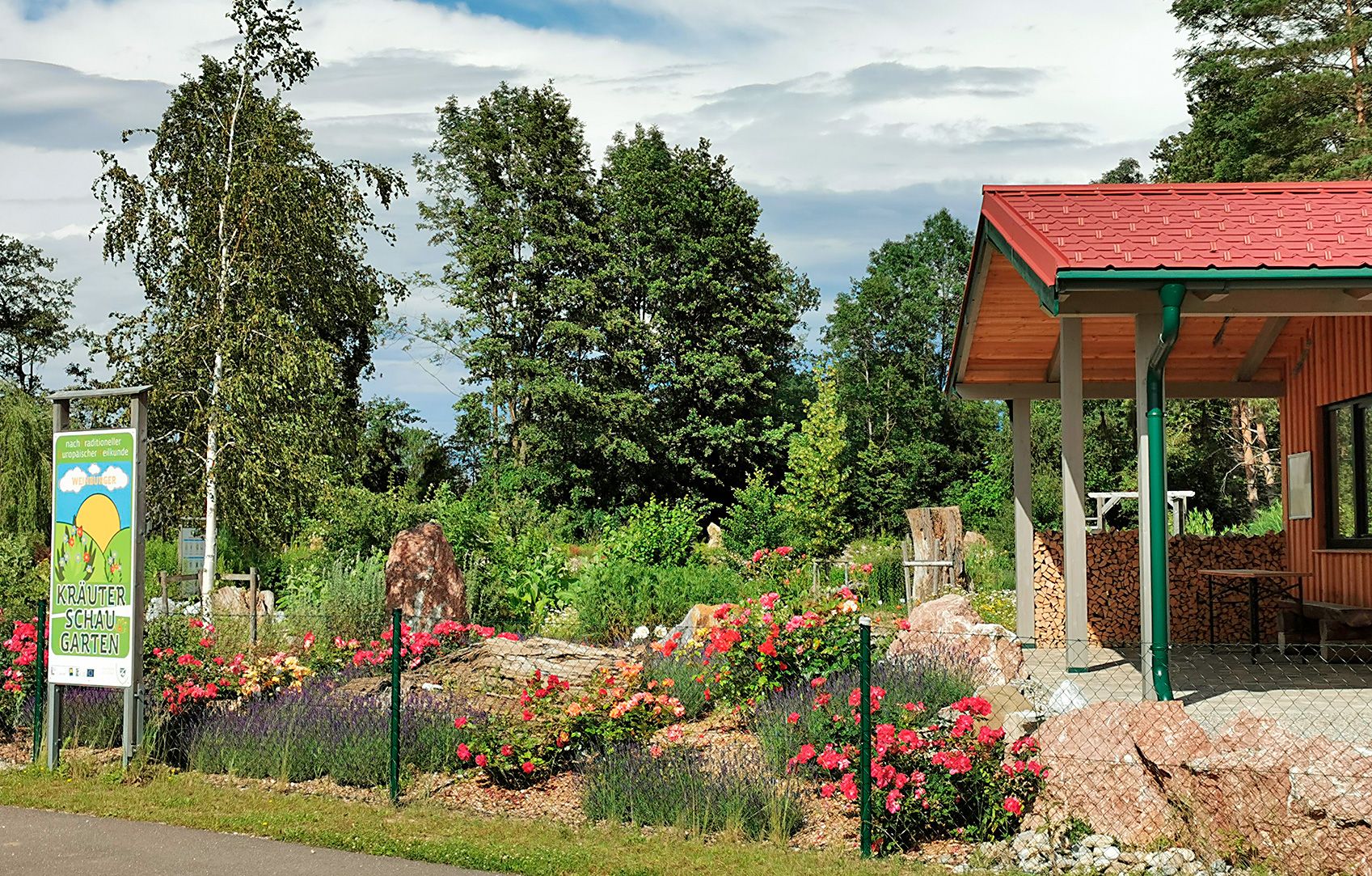 Ein Kräuterschaugarten mit bunten Blumen, Bäumen und einem Gebäude mit rotem Dach.