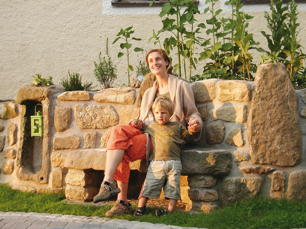 Eine Frau und ein Kind sitzen auf einer Steinbank vor einem mit Pflanzen bewachsenen Steingarten.