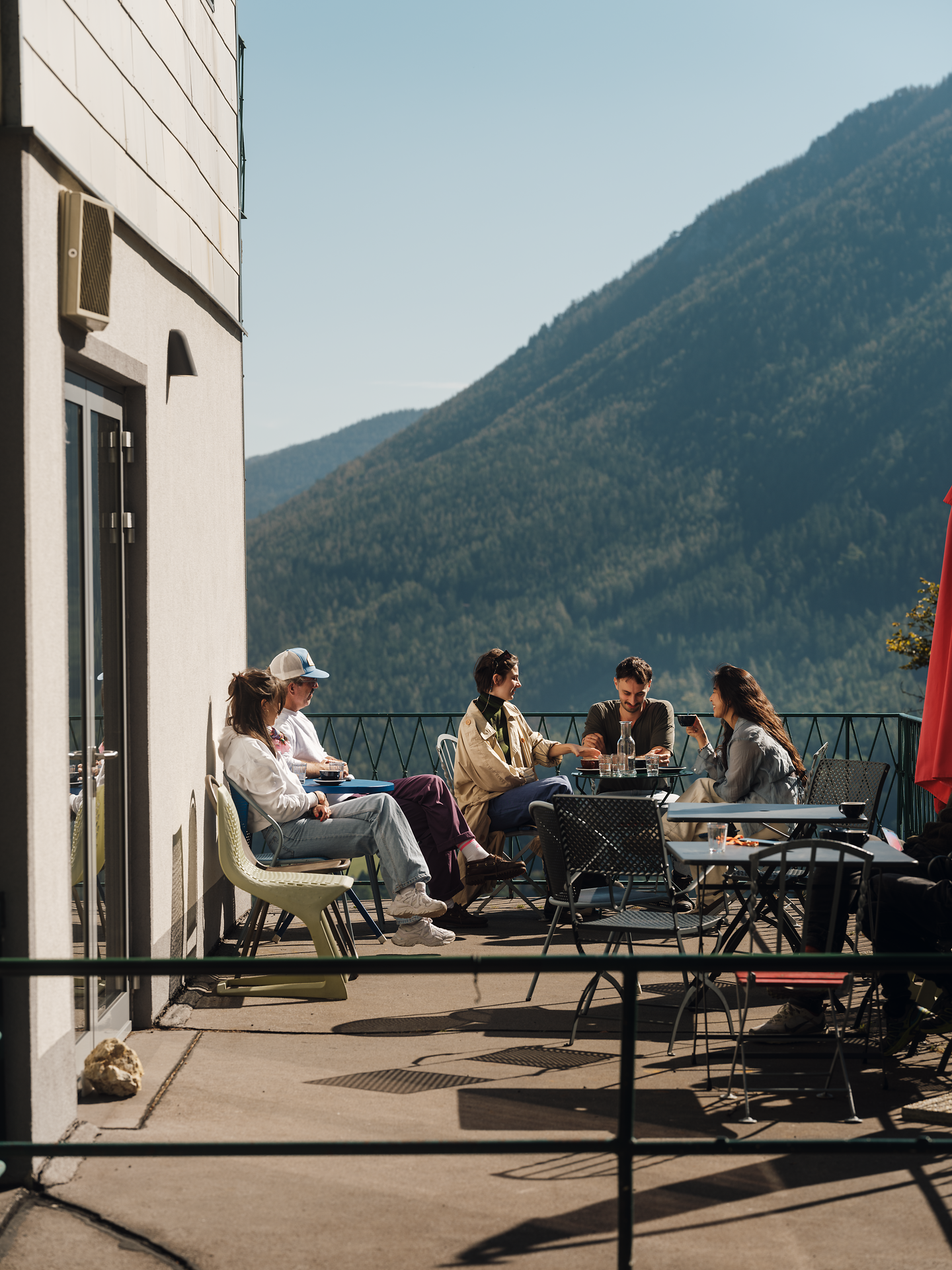 An einem sonnigen Tag genießen Freunde die entspannte Atmosphäre auf der Terrasse, umgeben von majestätischen Bergen. Die sanfte Brise und der Blick auf die atemberaubende Landschaft laden dazu ein, die Seele baumeln zu lassen und die Schönheit der Natur zu erleben.
