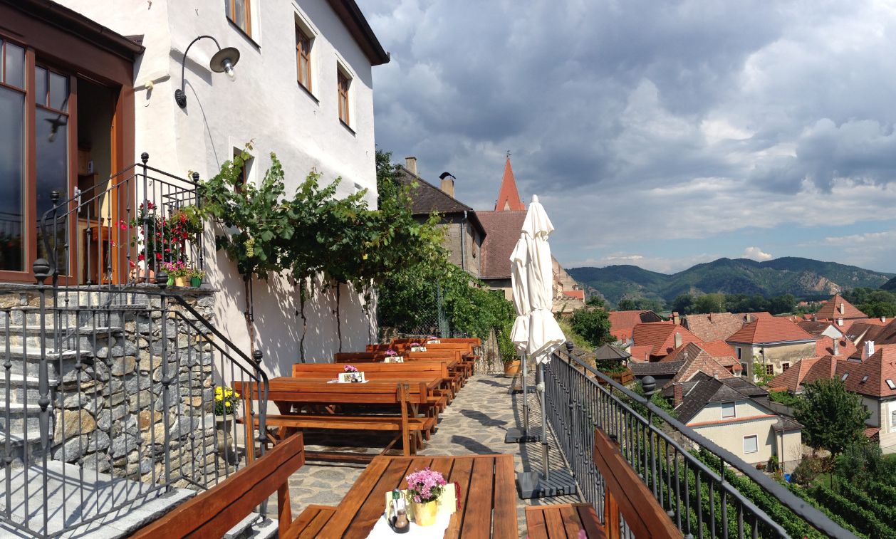 Terrasse mit Holzbänken und Tischen, Blick auf Dächer und Berge im Hintergrund.