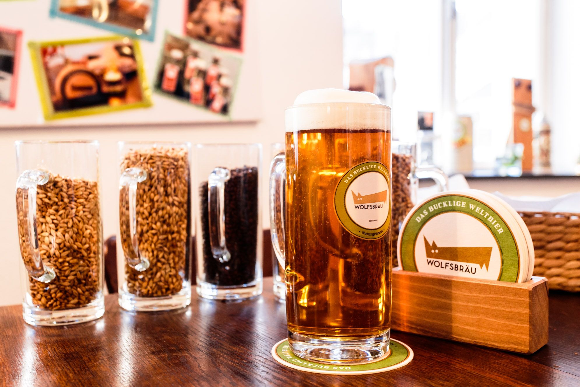 Krug mit Bier neben Bierdeckeln, im Hintergurnd Biergläser mit Getreide.