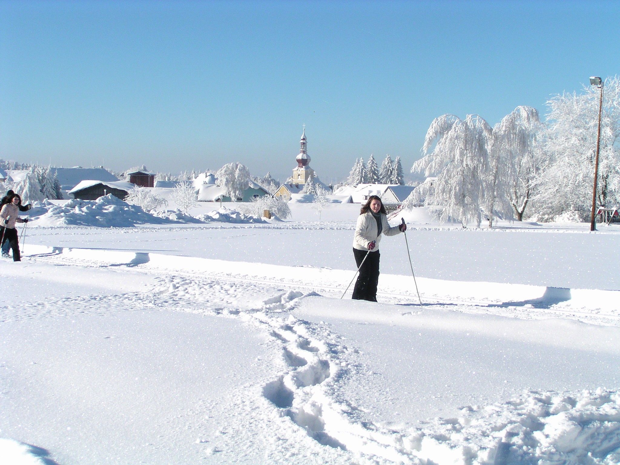 Zwei Personen beim Langlaufen in einer verschneiten Landschaft mit einer Kirche im Hintergrund.