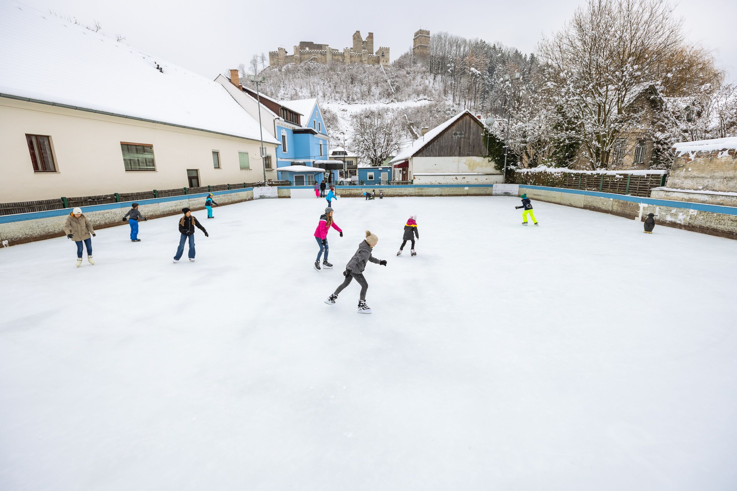 Menschen eislaufend auf einem Platz in Kirchschlag, im Hintergrund auf einem Hügel eine Burgruine.