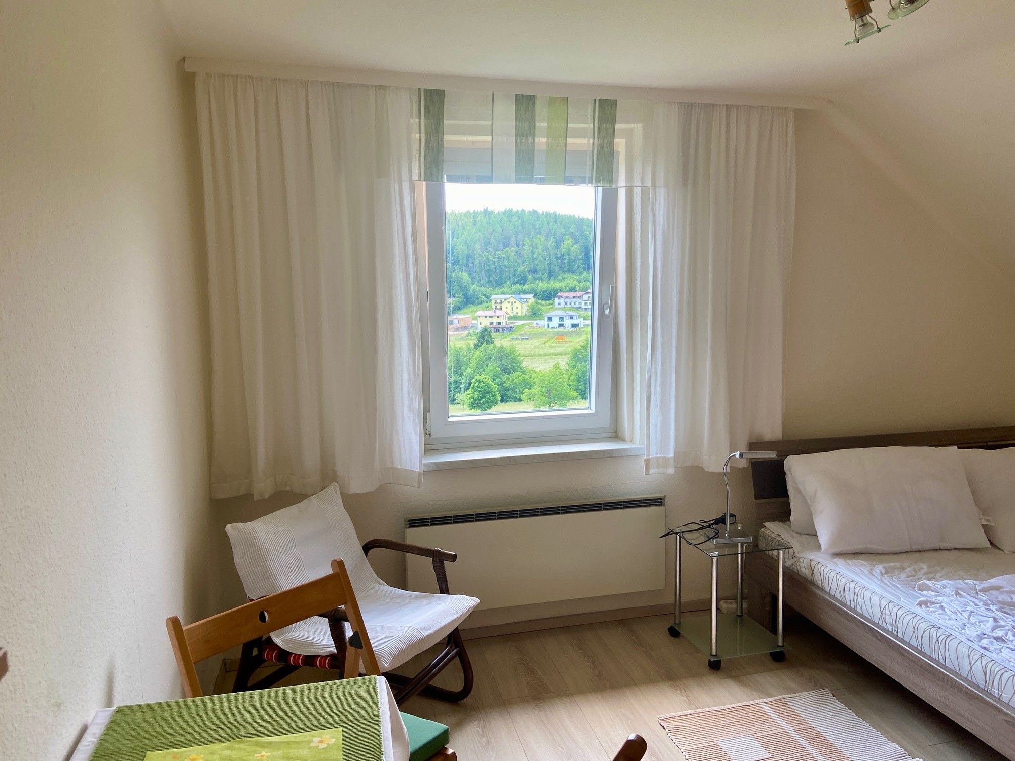 Helles Schlafzimmer mit Fensterblick auf grüne Landschaft, Bett, Stuhl und Tisch.