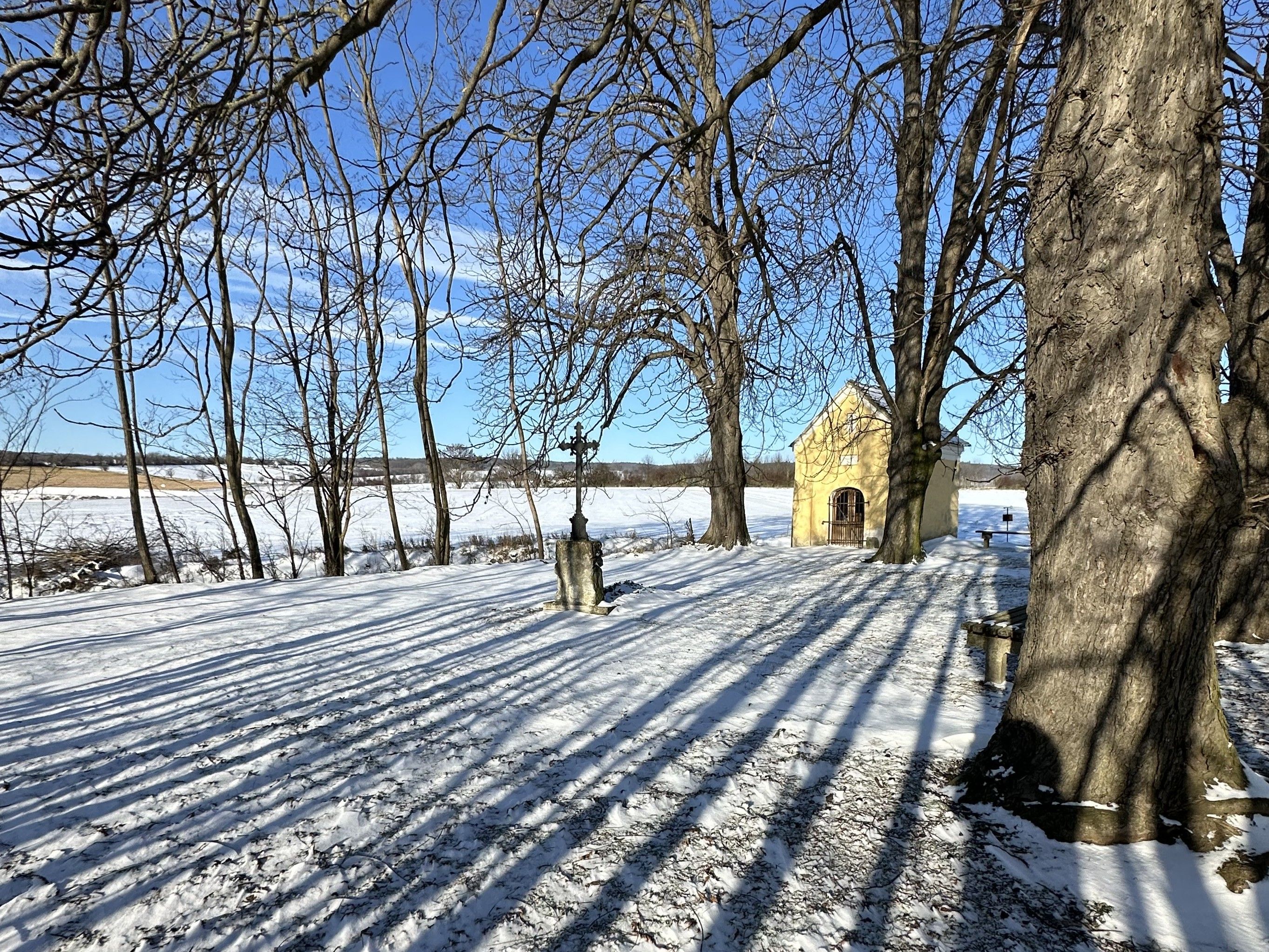 Winterlandschaft mit einer kleinen gelben Kapelle im Hintergrund, umgeben von schneebedeckten Bäumen und Feldern.