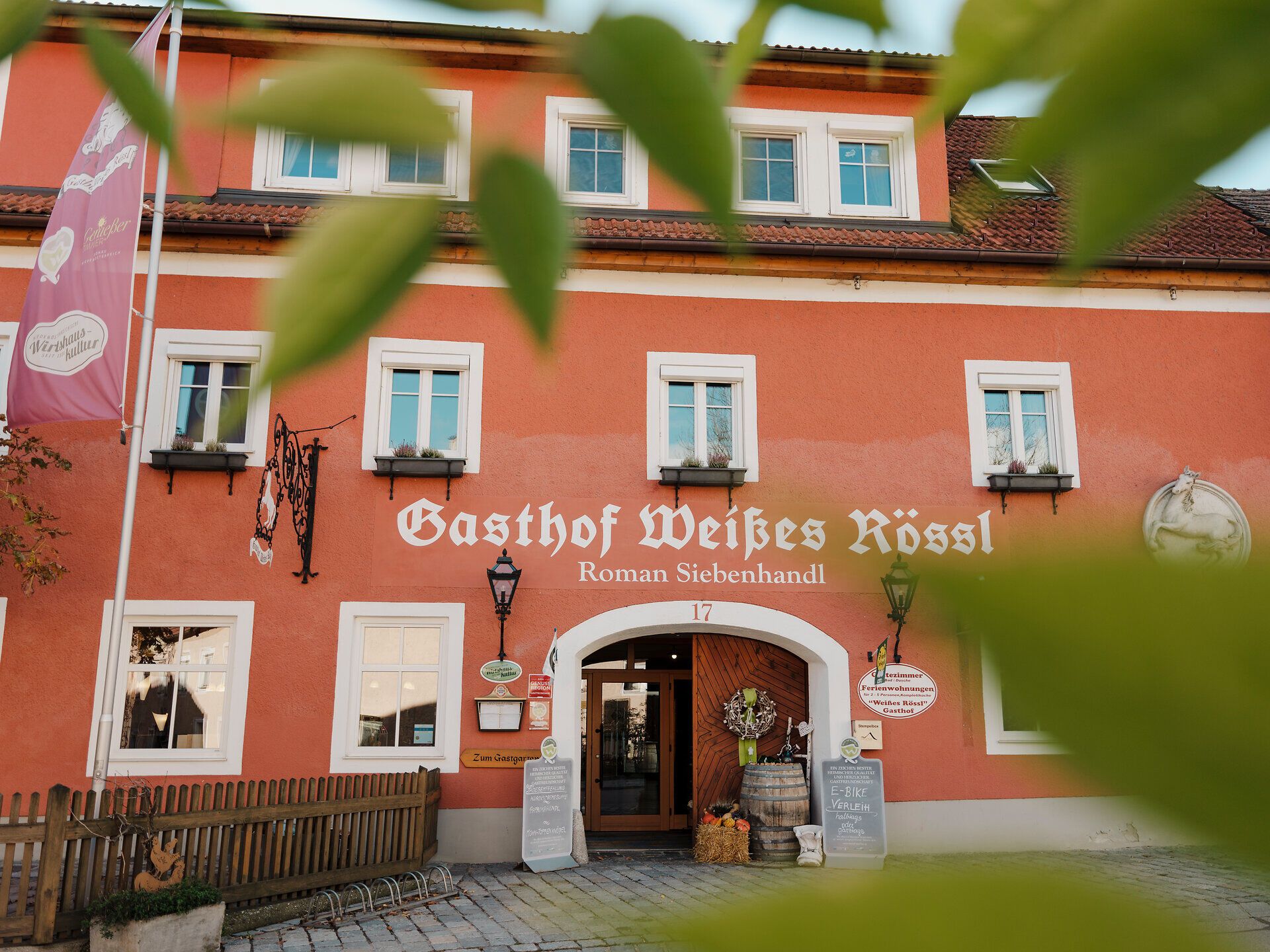 Inmitten der malerischen Landschaft Niederösterreichs strahlt der Gasthof Weißes Rössl eine einladende Gemütlichkeit aus. Die warmen Farben der Fassade harmonieren perfekt mit der umgebenden Natur, während die blühenden Pflanzen im Vordergrund den Sommerzauber unterstreichen. Hier erleben Gäste die traditionelle Wirtshauskultur und die herzliche Gastfreundschaft der Region.