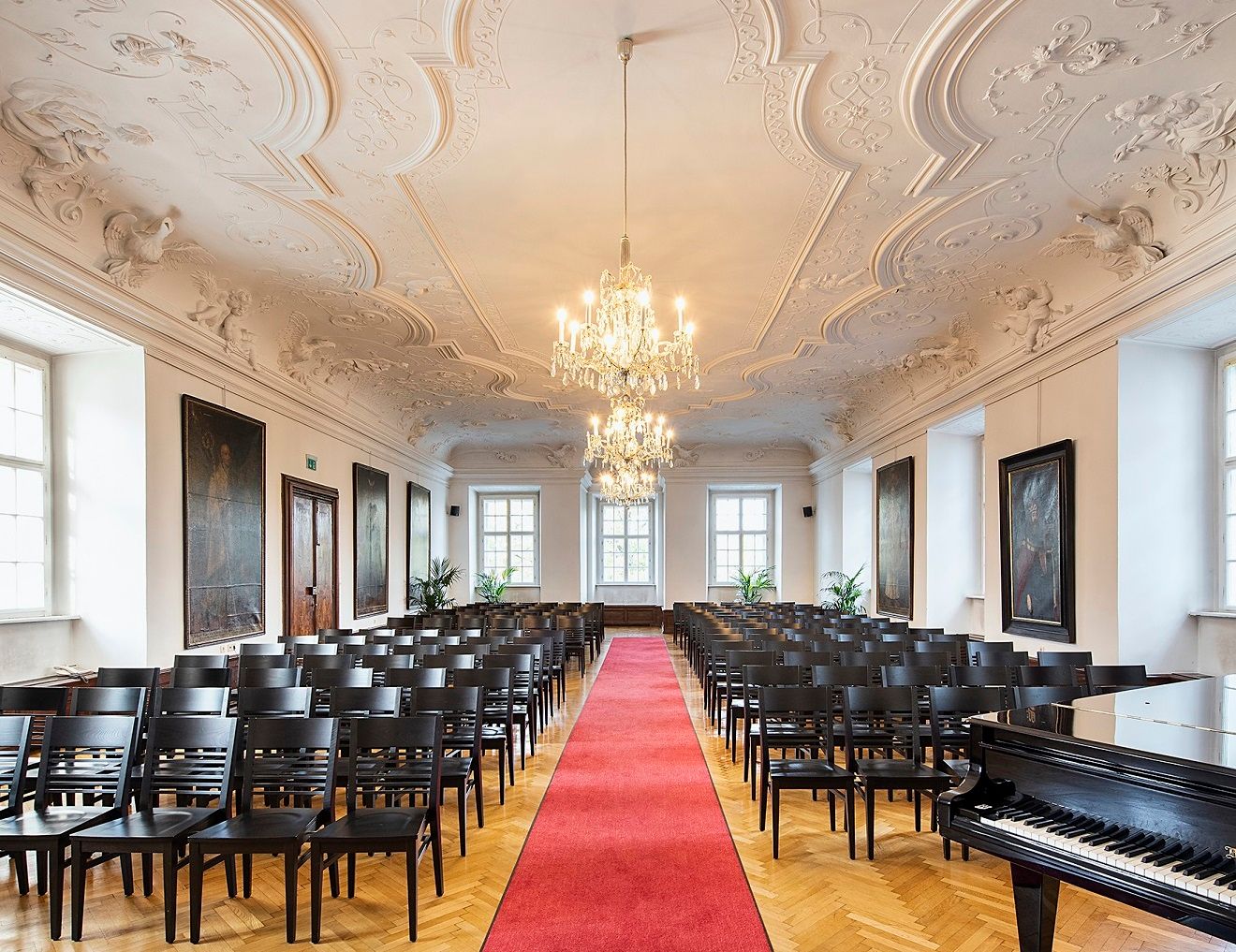 Eleganter Saal mit Stuckdecke, Kronleuchtern, Stuhlreihen und rotem Teppich.