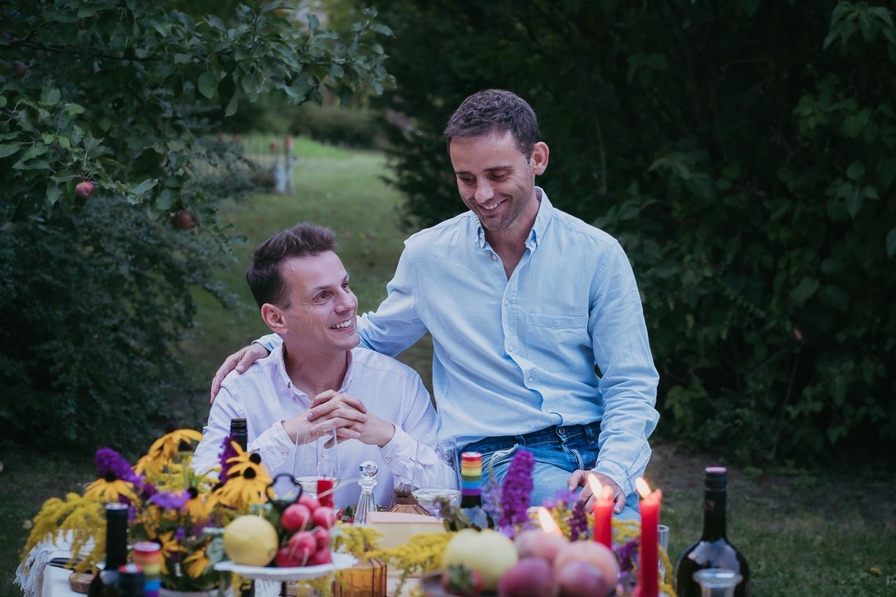 Zwei Männer sitzen an einem Tisch im Freien, umgeben von Blumen und Kerzen.