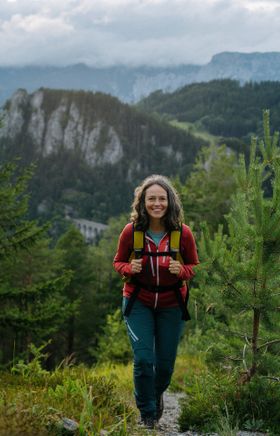 Irene Beckmann steht vor einer malerischen Hügelkulisse mit einem traditionellen Gebäude im Hintergrund. Nebelschwaden und bewaldete Berge vervollständigen die idyllische Szene, die Freude an der Natur und kultureller Entdeckung vermittelt.