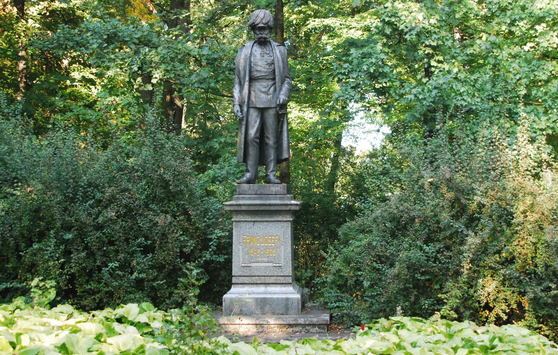 Statue von Kaiser Franz Joseph I. in einem Park, umgeben von Bäumen und Sträuchern.