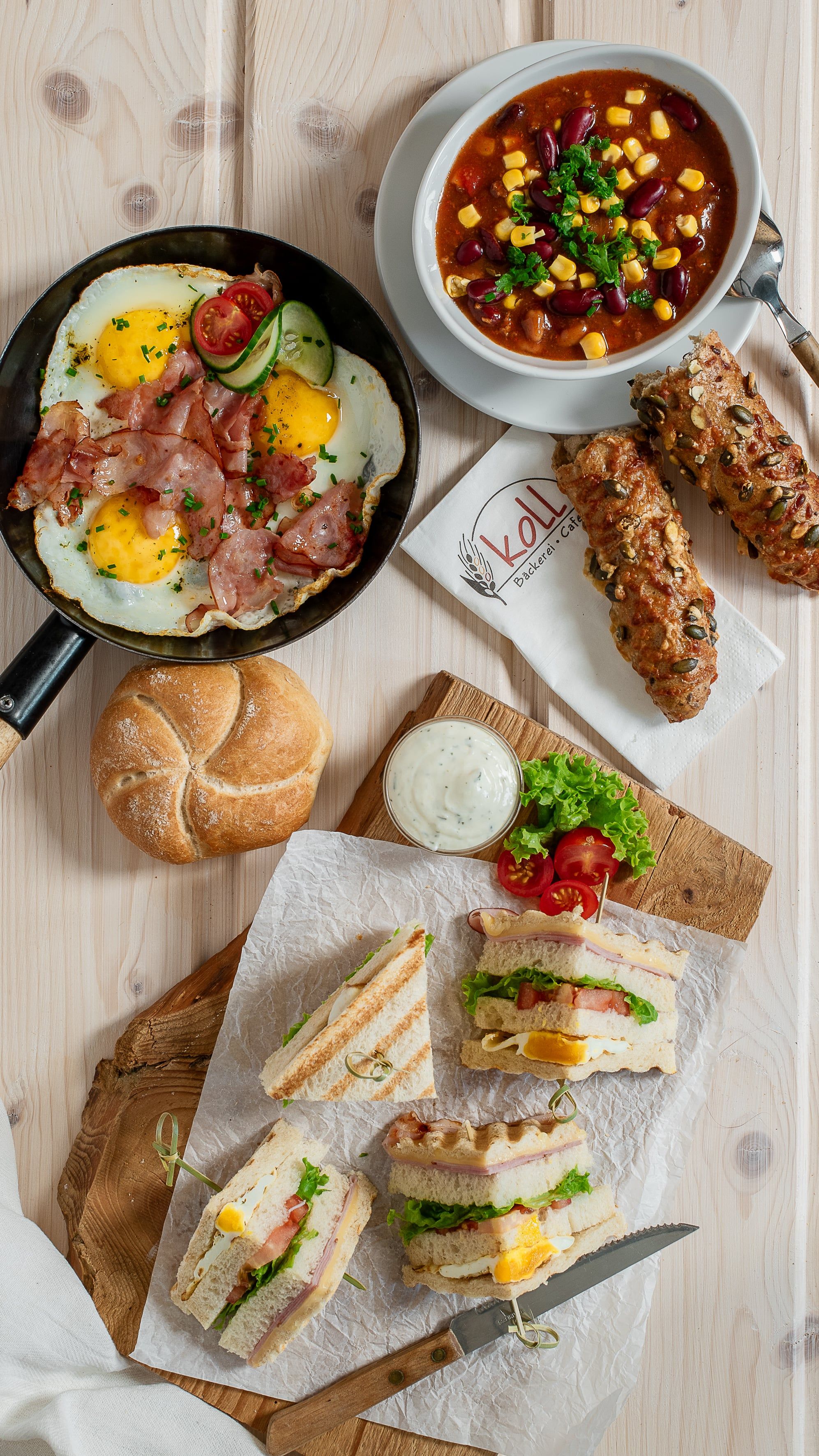 Verschiedene warme Snacks auf einem Tisch, darunter Spiegeleier mit Speck, Chili con Carne, belegte Brote und Brötchen.
