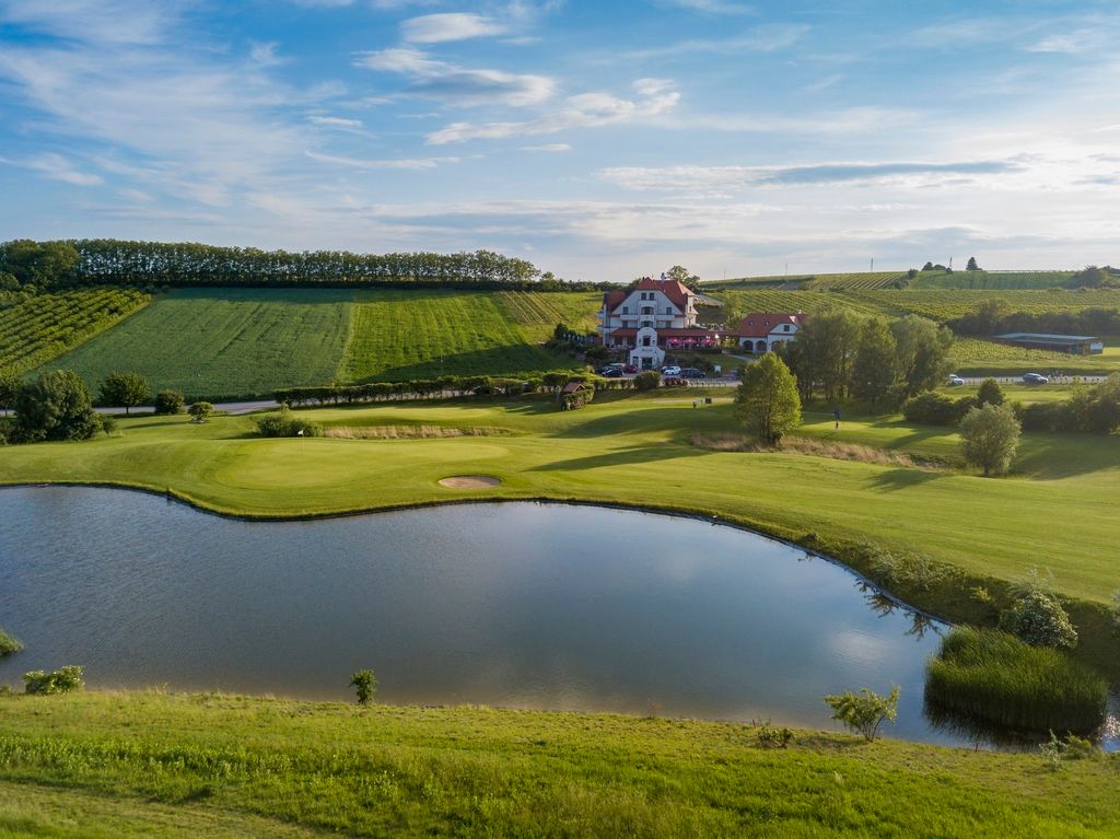 Landschaftsaufnahme eines Golfplatzes mit Teich und Clubhaus im Hintergrund.