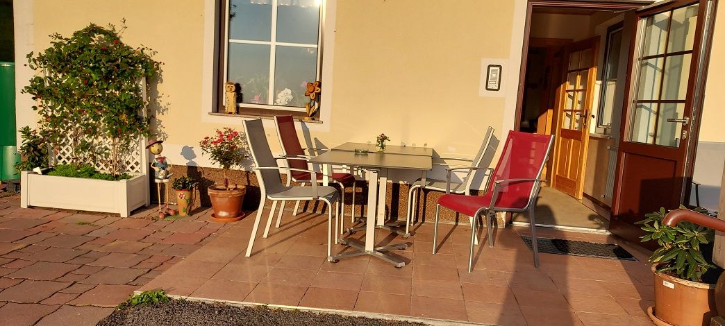 Terrasse mit Tisch und Stühlen vor einem Gebäude, umgeben von Pflanzen.