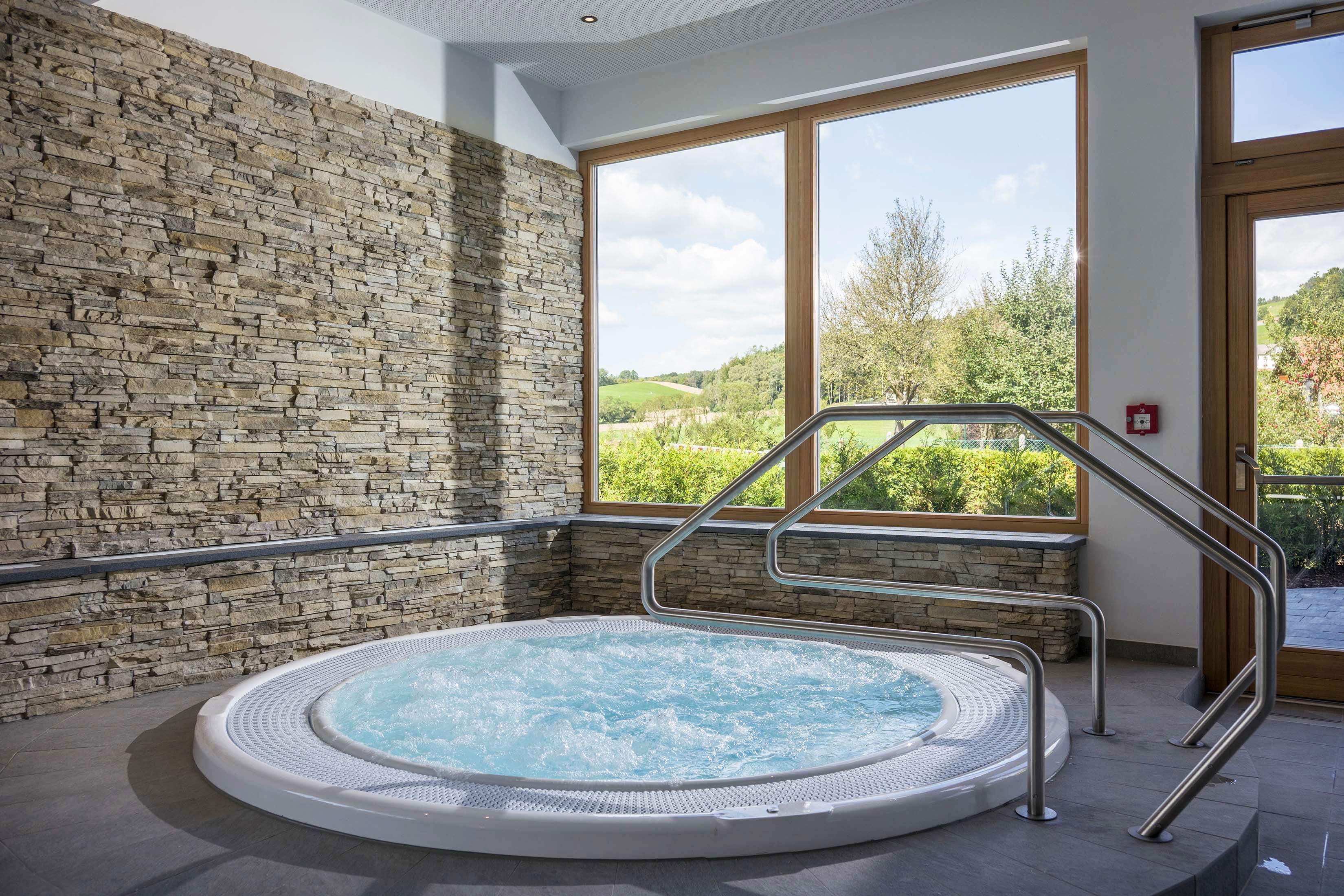 Wellnessraum mit Whirlpool, Steinwänden und großem Fenster mit Blick ins Grüne.