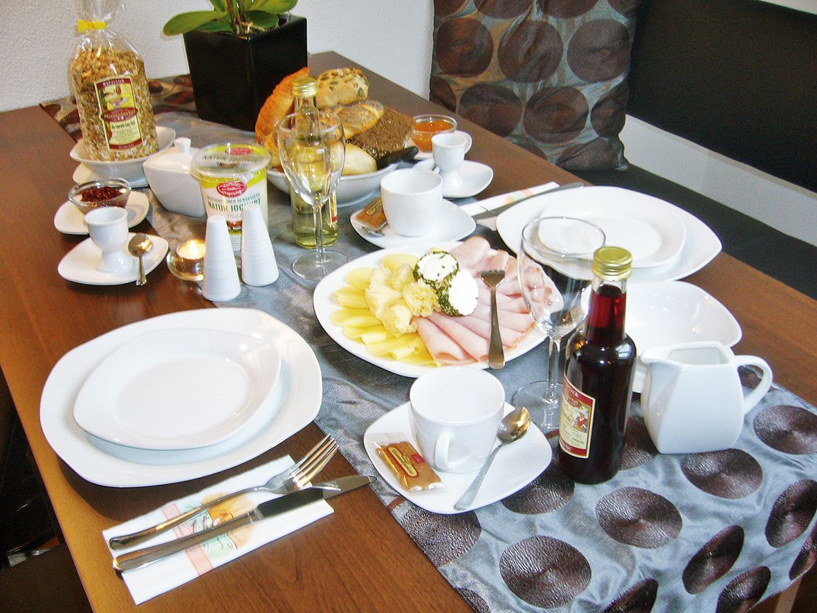 Frühstückstisch mit Geschirr, Aufschnitt, Brot, Müsli und Getränken.