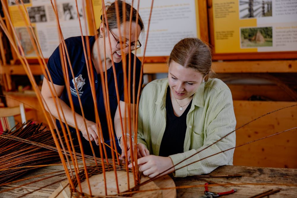 Zwei Personen flechten gemeinsam einen Korb aus Weidenruten in einer Werkstatt.