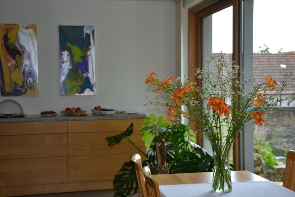 Ein Frühstücksraum mit einem Tisch, auf dem ein Blumenstrauß steht. Im Hintergrund sind Kunstwerke an der Wand und eine Anrichte mit Obst.