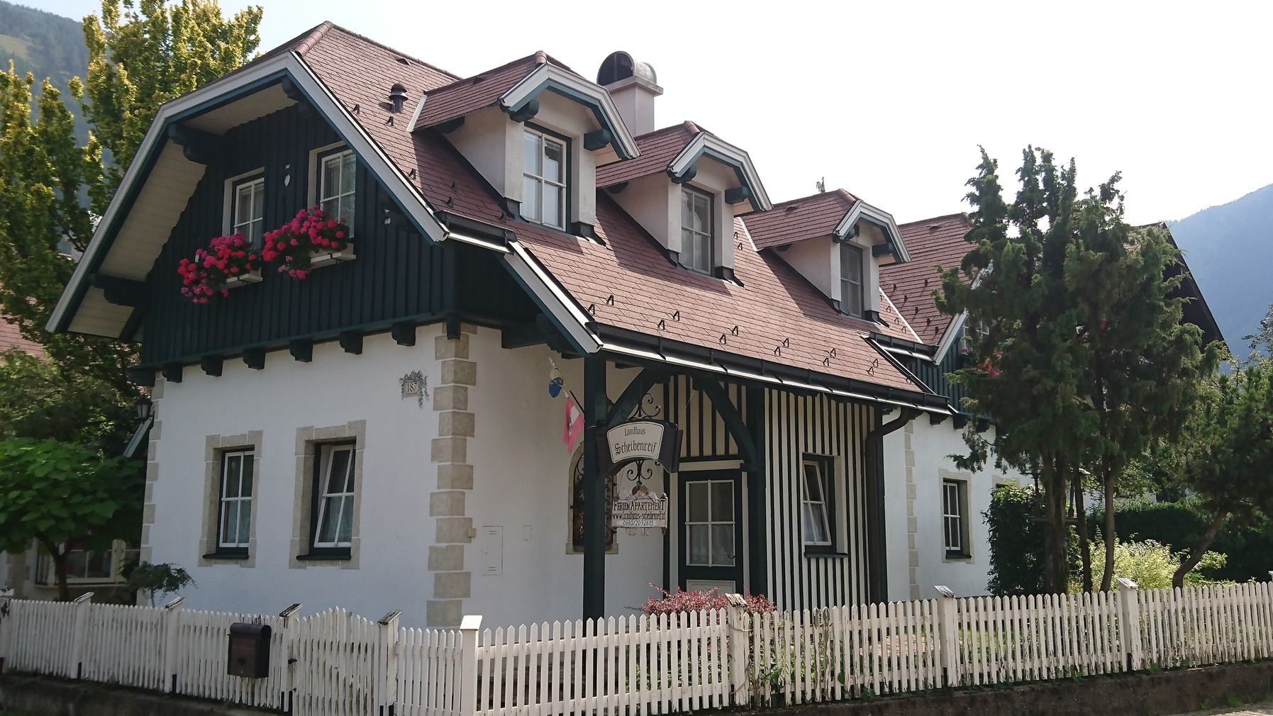 Ein traditionelles Landhaus mit Holzverkleidung, roten Dachziegeln und Blumenkästen vor den Fenstern. Ein weißer Holzzaun umgibt das Grundstück.