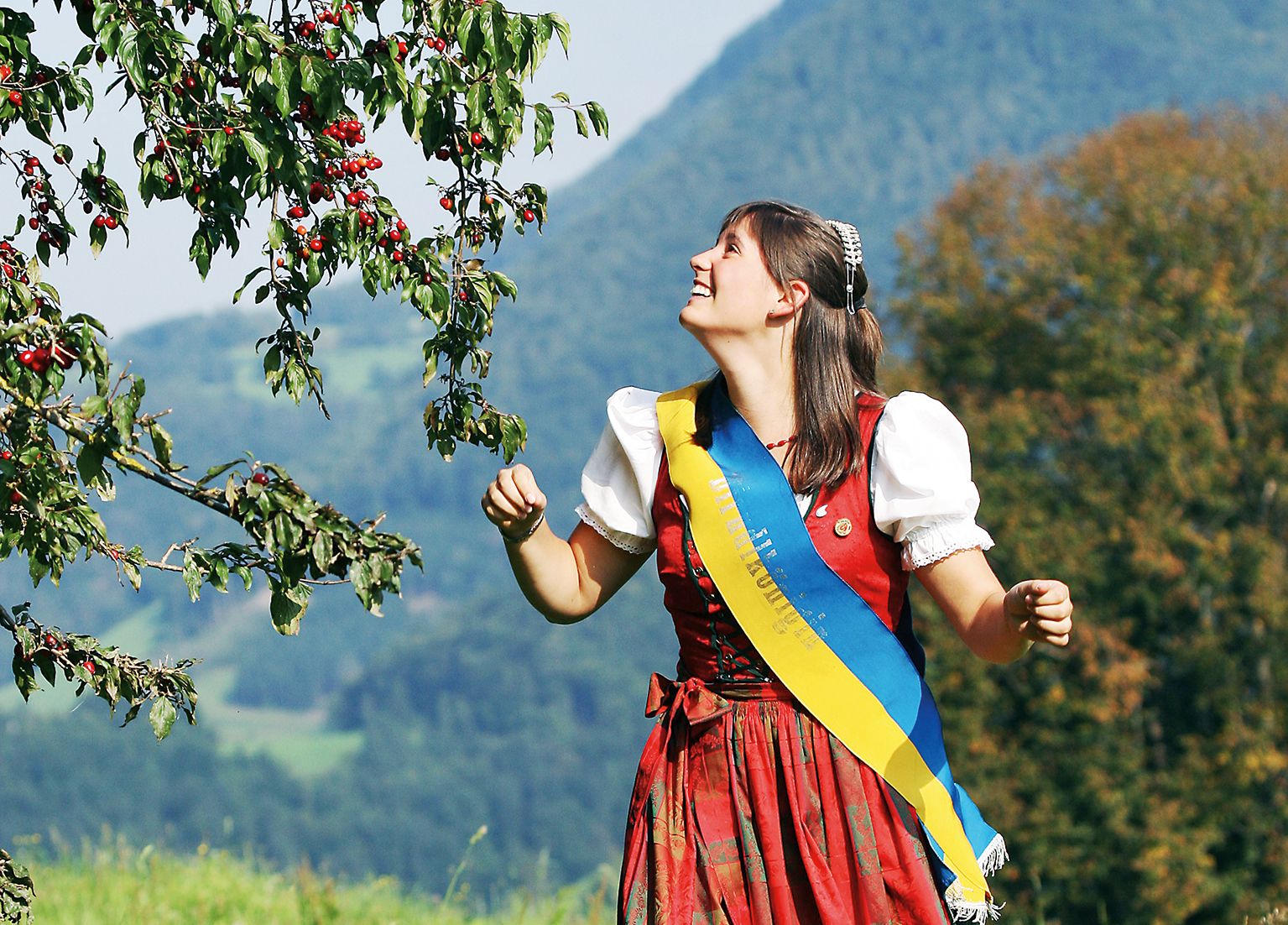 Frau in traditioneller Tracht steht unter einem Baum mit roten Früchten in einer ländlichen Landschaft.