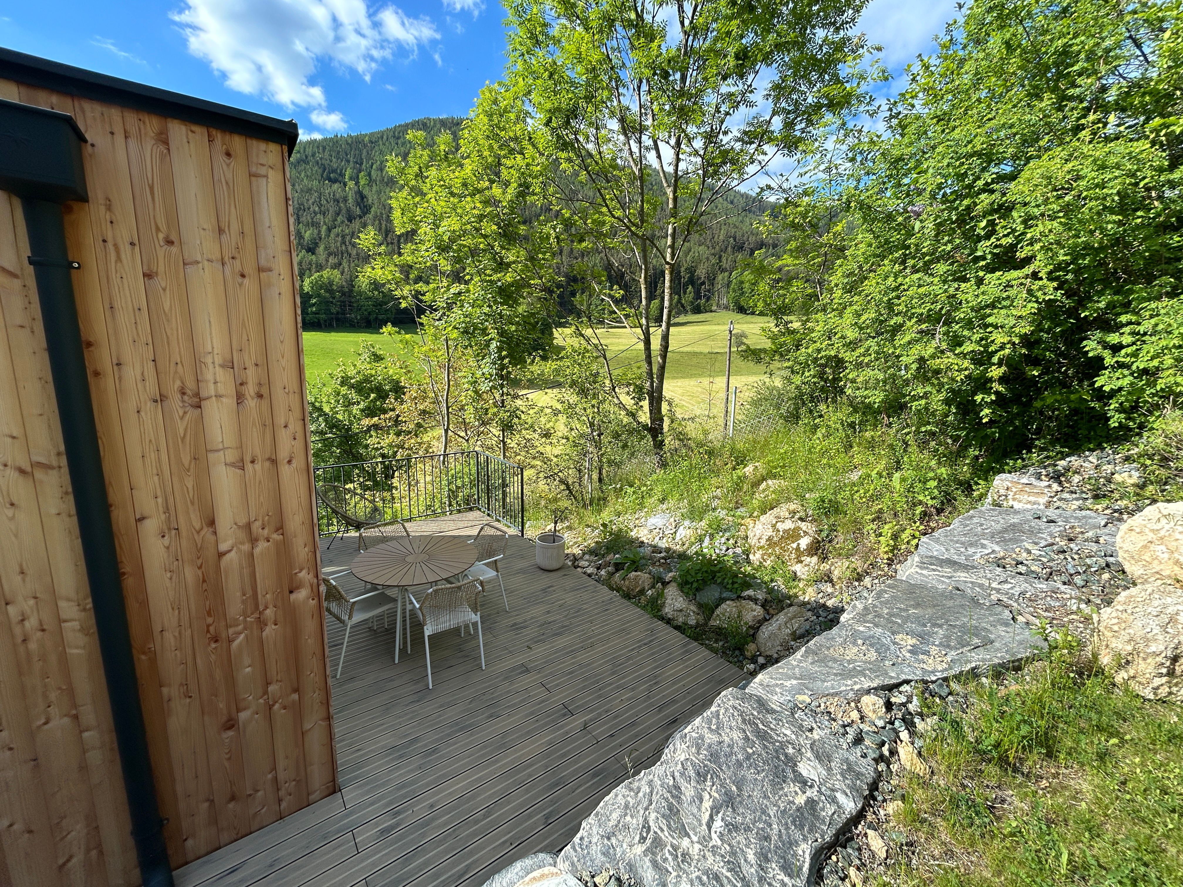 Terrasse mit Holztisch und Stühlen, umgeben von Bäumen und Wiese, mit Blick auf bewaldete Hügel.