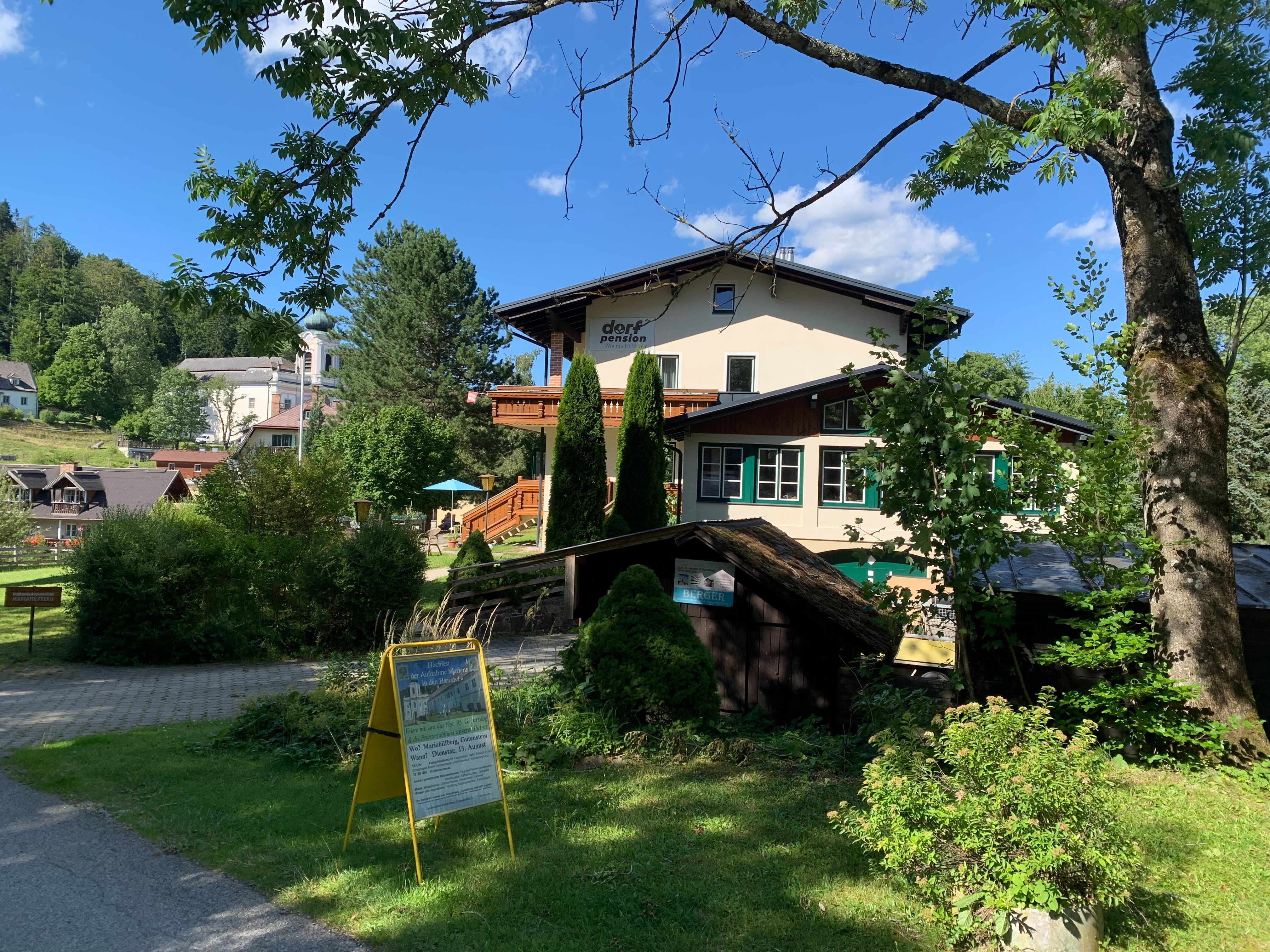 Eine Dorfpension in einer grünen, ländlichen Umgebung mit Bäumen und einem blauen Himmel.