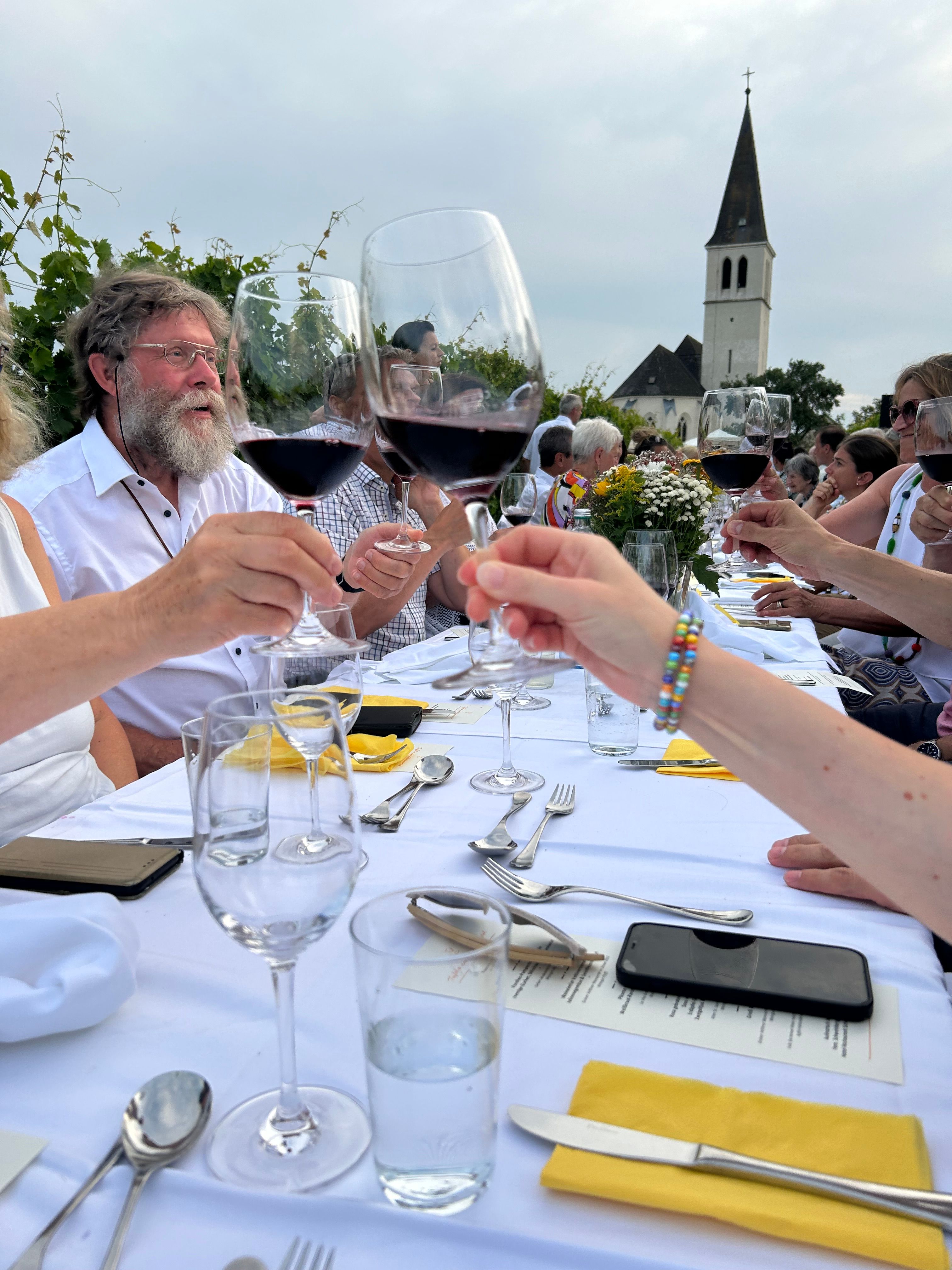 Menschen stoßen mit Weingläsern an einem langen Tisch im Freien an, im Hintergrund eine Kirche.