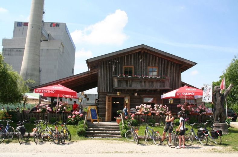 Holzhütte mit Terrasse, Fahrrädern und Sonnenschirmen vor einem Industriegebäude.